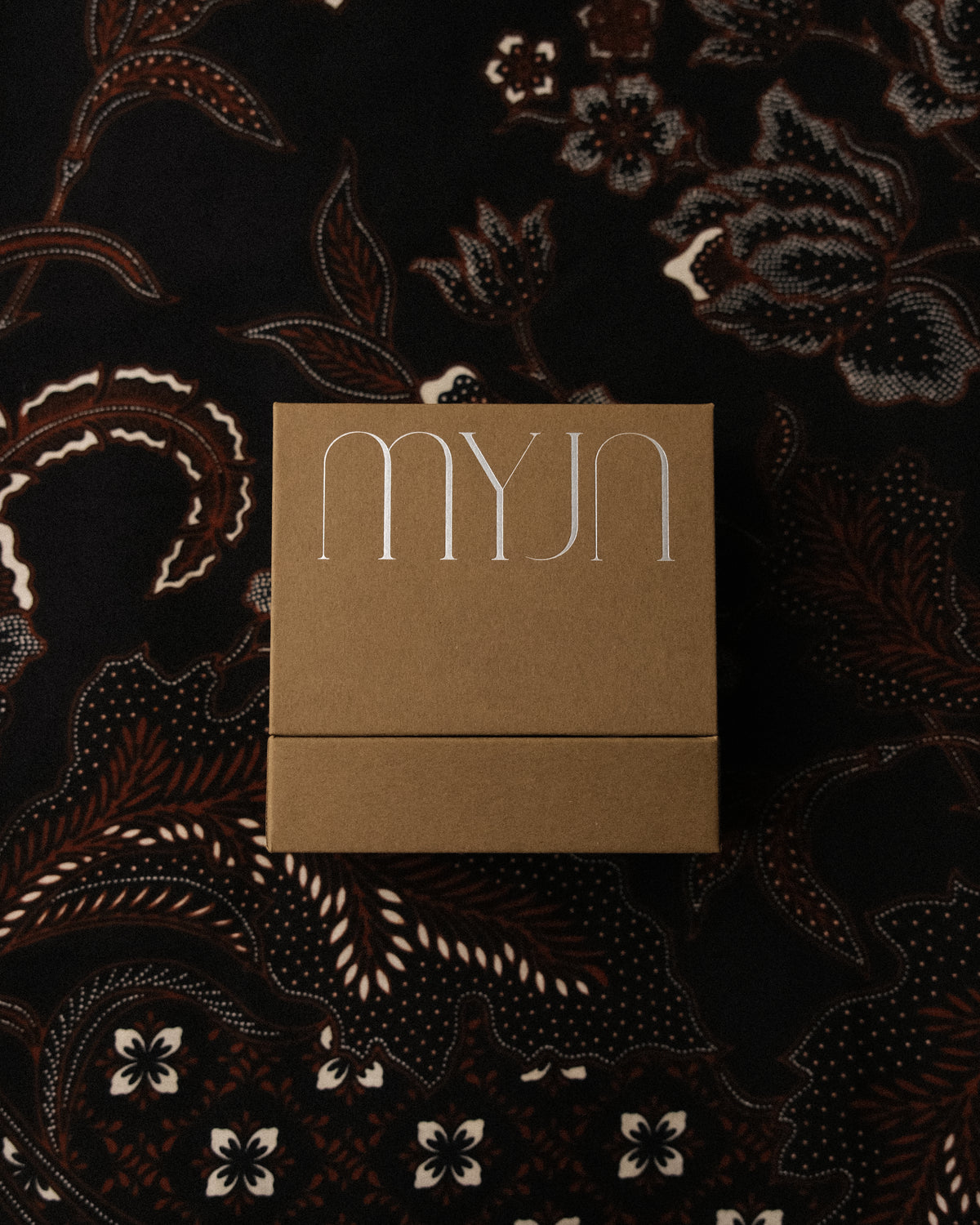 A square kraft paper box with the brand name 'myjn' in a stylized silver font, placed on a dark patterned fabric background.