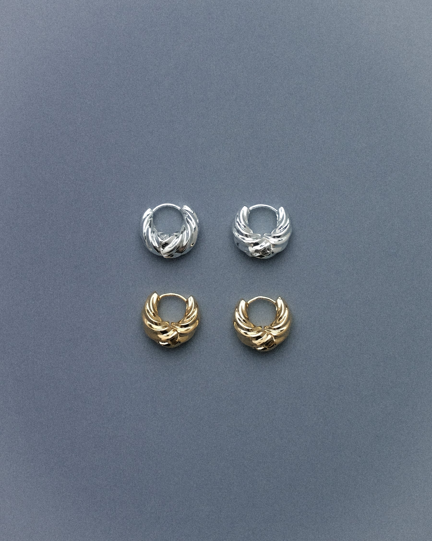 A pair of silver plated brass spiral hoop earrings and a pair of gold plated brass spiral hoop earrings, captured against a muted grey blue background, highlighting their sculptural spiral form with a clean, minimal aesthetic.