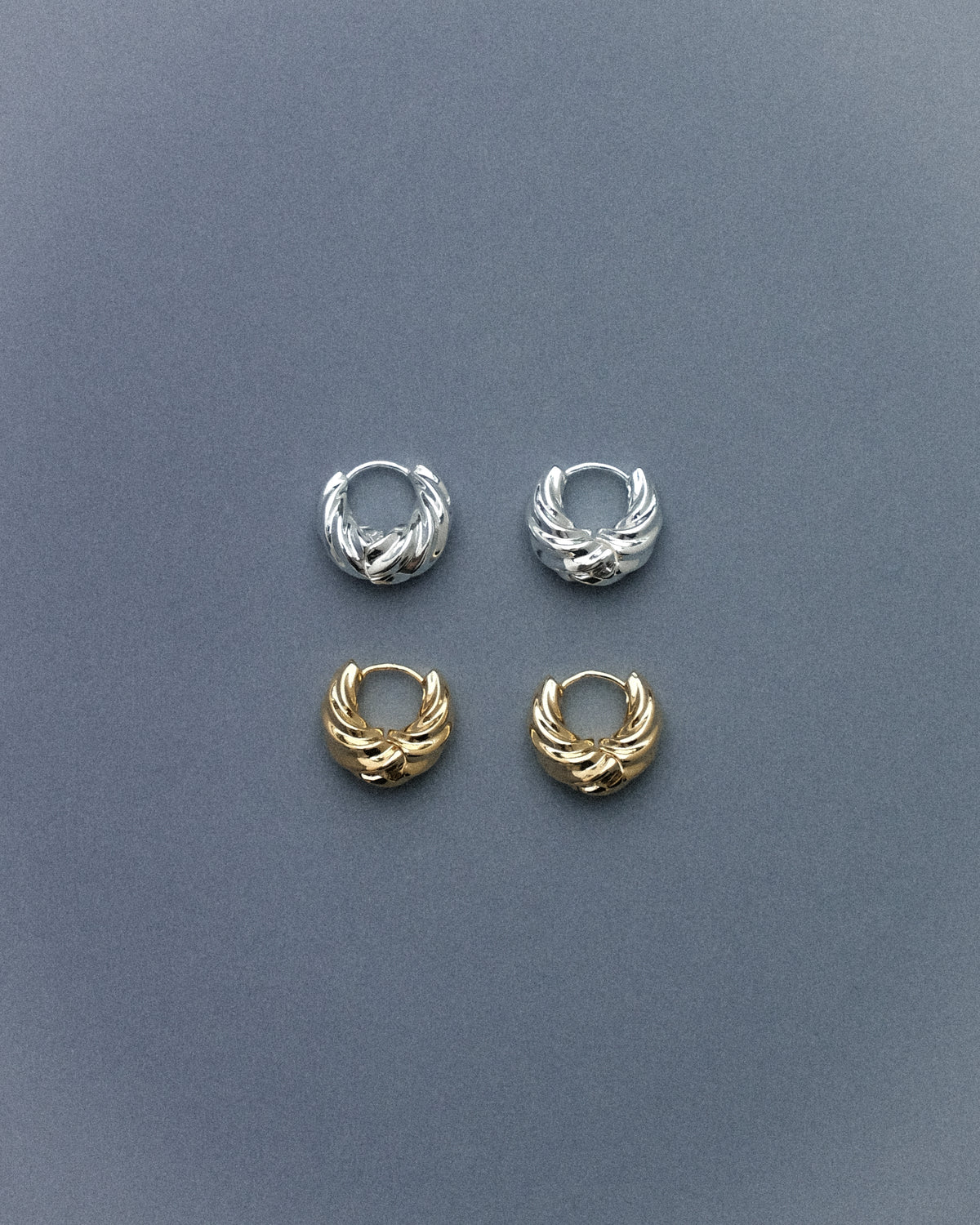 A pair of silver plated brass spiral hoop earrings and a pair of gold plated brass spiral hoop earrings, captured against a muted grey blue background, highlighting their sculptural spiral form with a clean, minimal aesthetic.