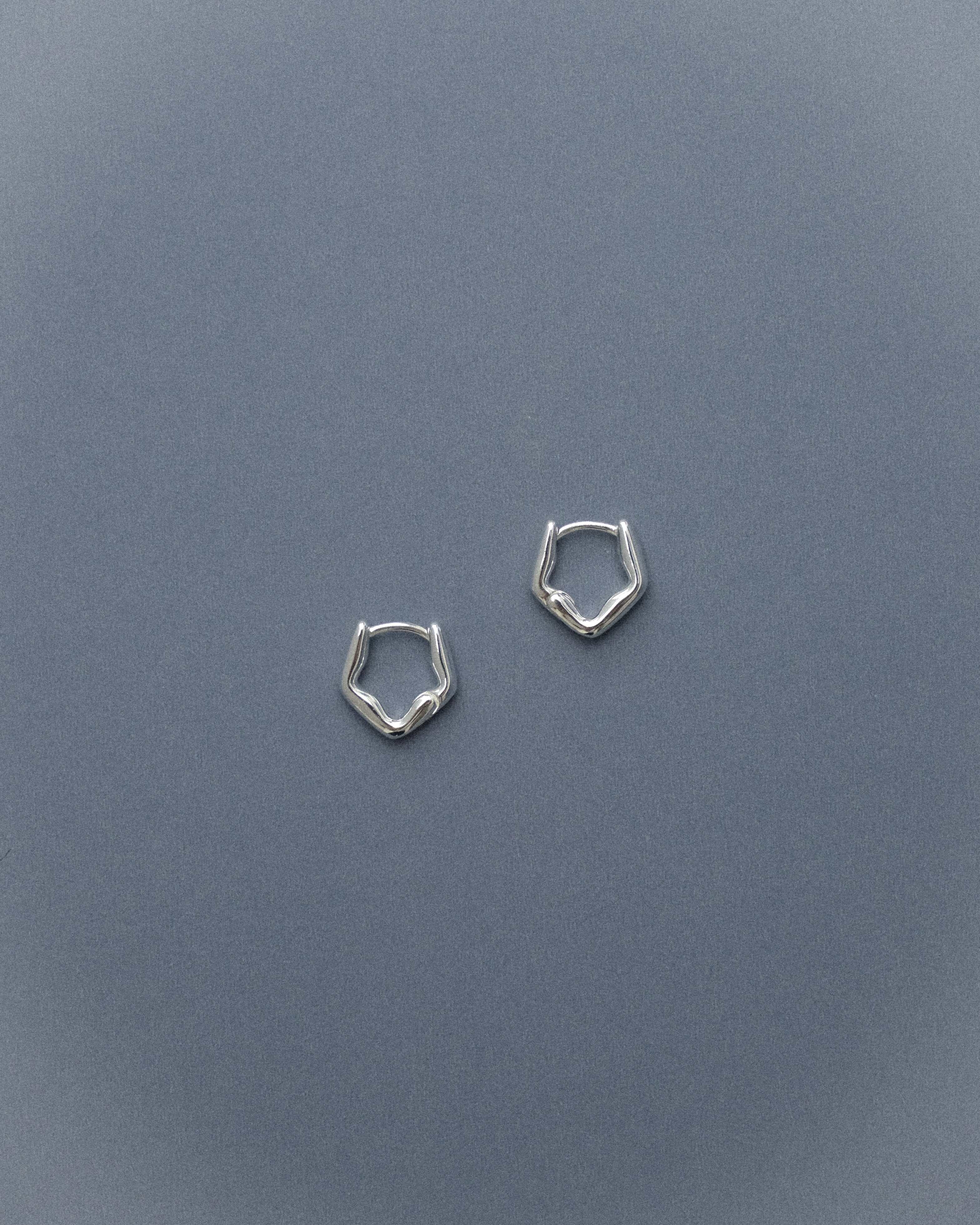 A pair of sculptural silver earrings set against a soft grey-blue background, their fluid forms quietly catching the light.