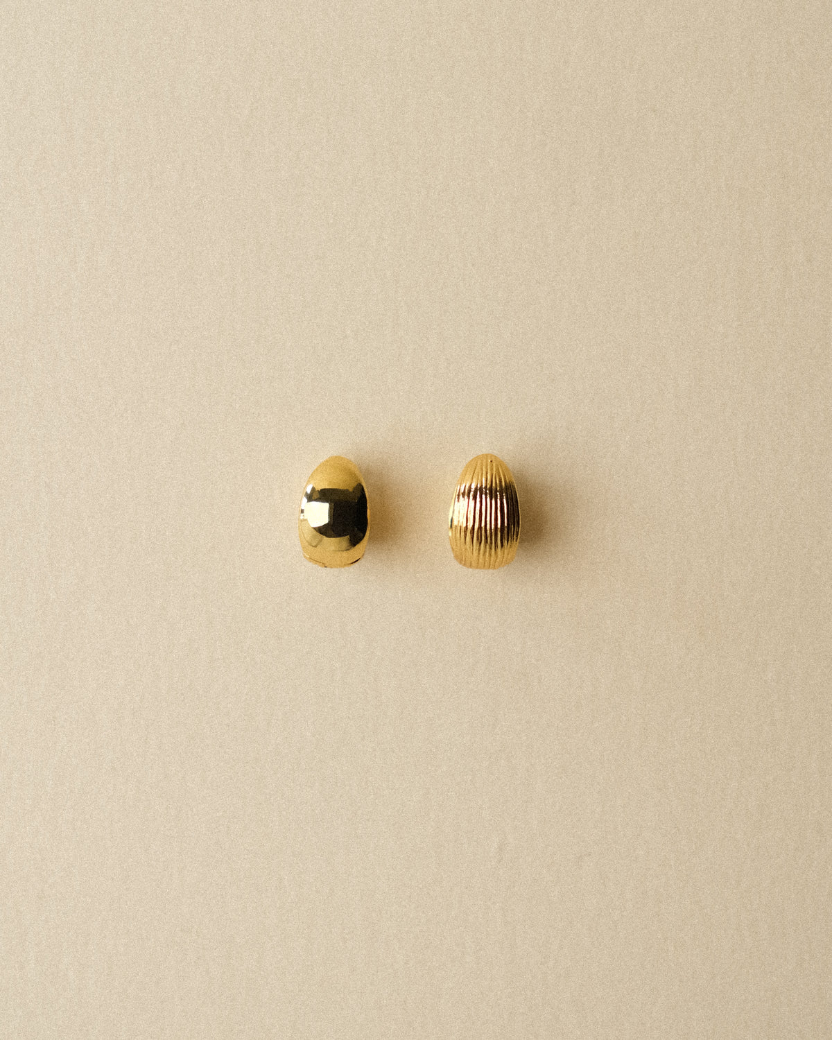 A pair of small 18k gold-plated brass reversible hoop earrings, one smooth and one ribbed, displayed on a textured wabi-sabi warm background.