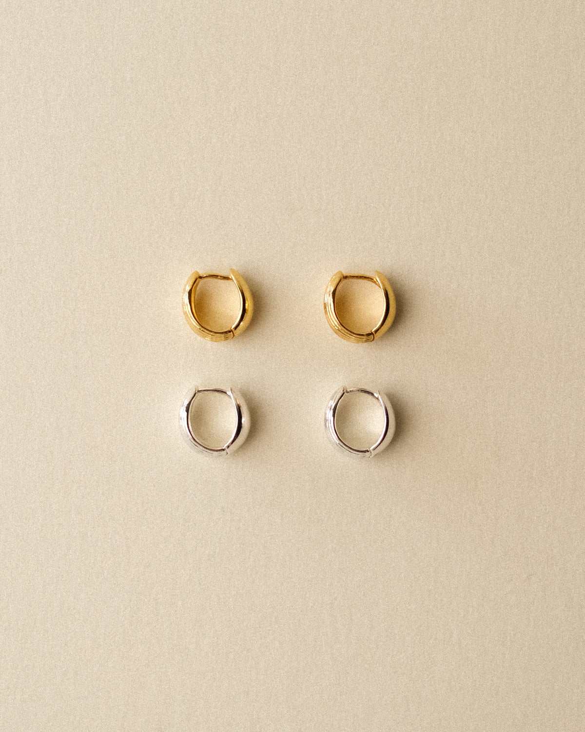 Four small reversible hoop earrings arranged in a 2x2 grid on a warm beige background. The top row features textured gold hoops, and the bottom row features textured silver hoops.