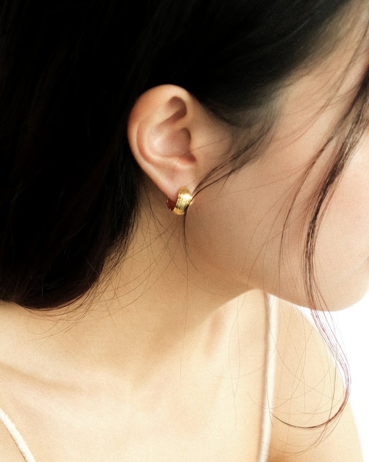 A close-up side profile of a woman wearing a small gold reversible hoop earring, shown against her ear and dark hair.