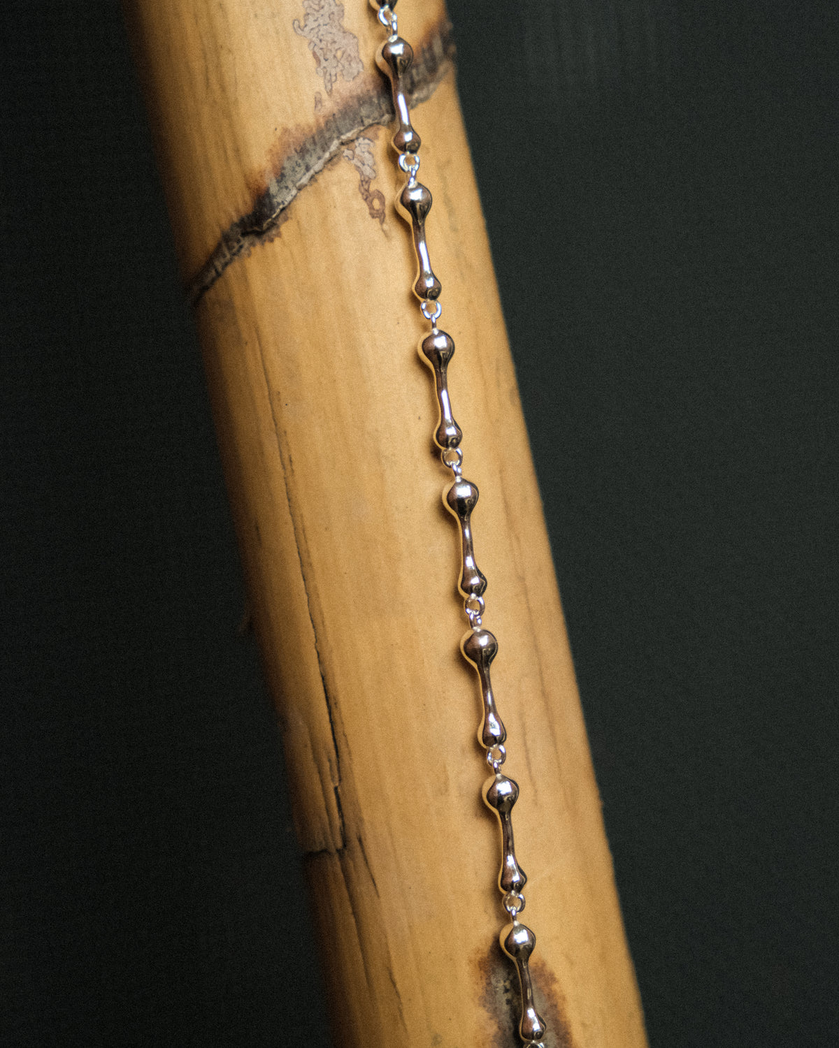 A close-up of a silver droplet link chain draped vertically over a bamboo cylinder, highlighting its delicate form and polished details against a deep, dark background.