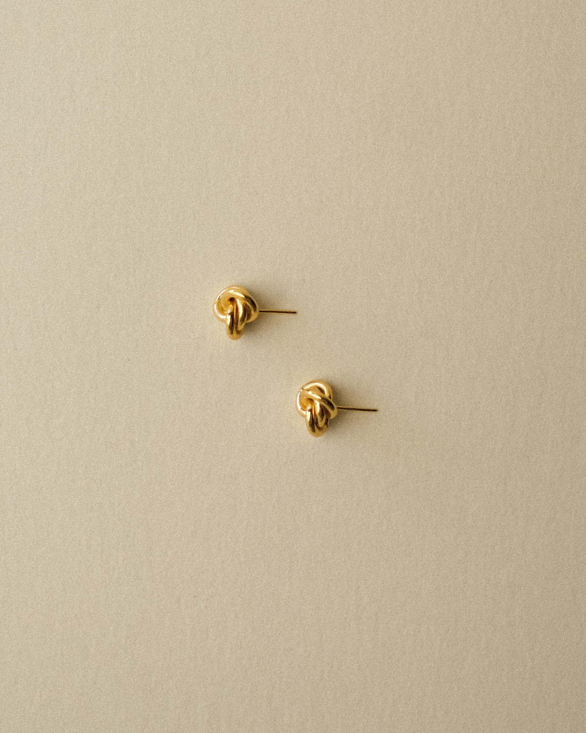 A pair of small gold knot stud earrings on a plain warm wabi-sabi background.