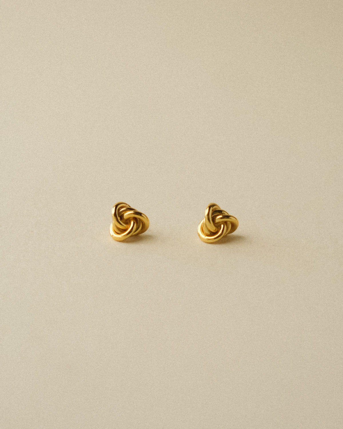 A pair of gold knot stud earrings on a plain warm wabi-sabi background.
