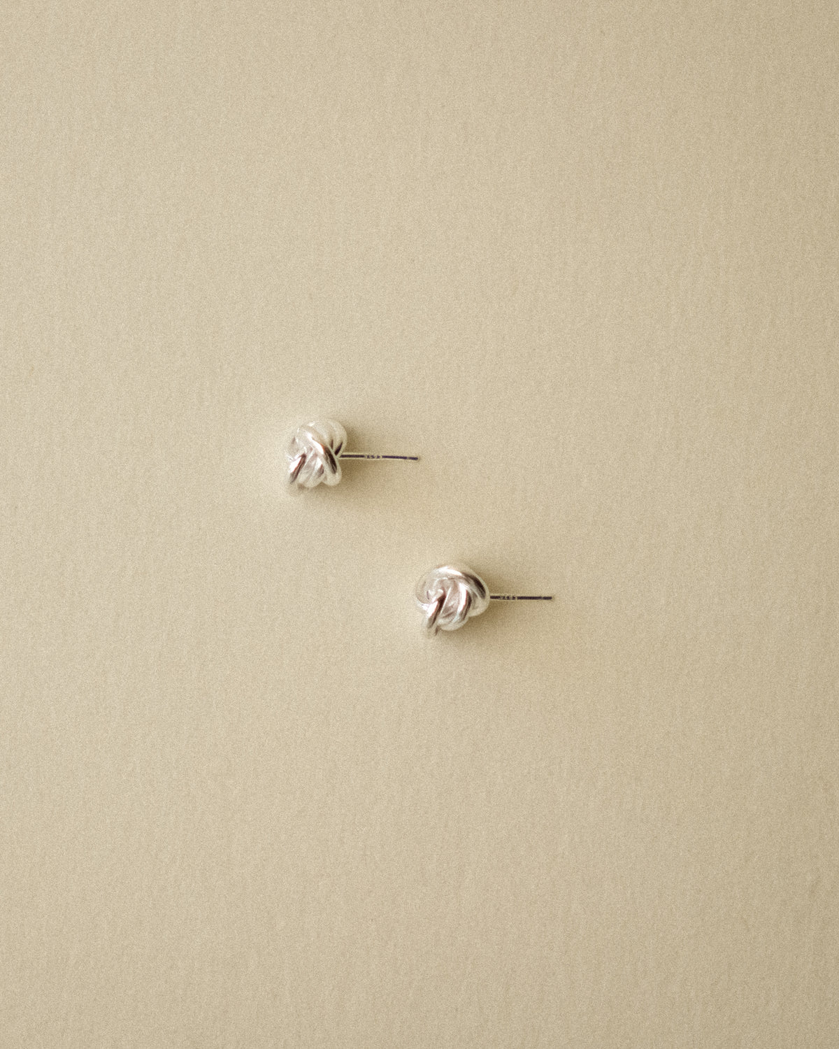 A pair of small silver knot stud earrings on a plain warm wabi-sabi background.