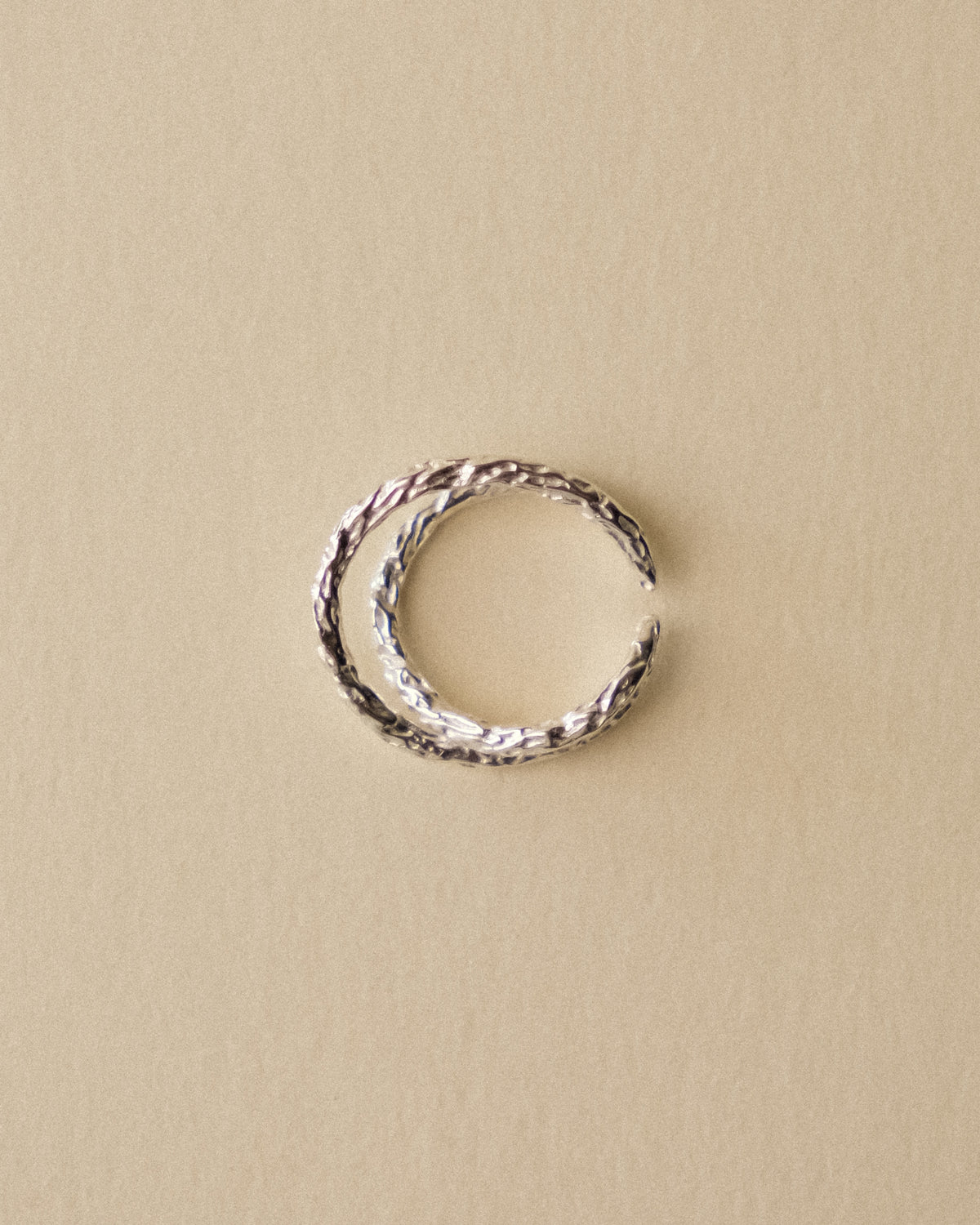 A hammered crossed ring laid flat, captured from above to showcase the complete form and sculptural silhouette against a warm, minimal wabi-sabi background.