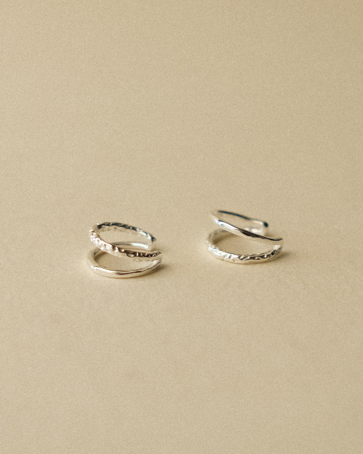A pair of silver ear cuffs embracing the ear with a double-layered design, balancing the raw texture of hand-hammered silver with the refined sheen of a polished finish, captured from the side with one ear cuff positioned upside down in a sculptural composition against a warm wabi-sabi inspired background.