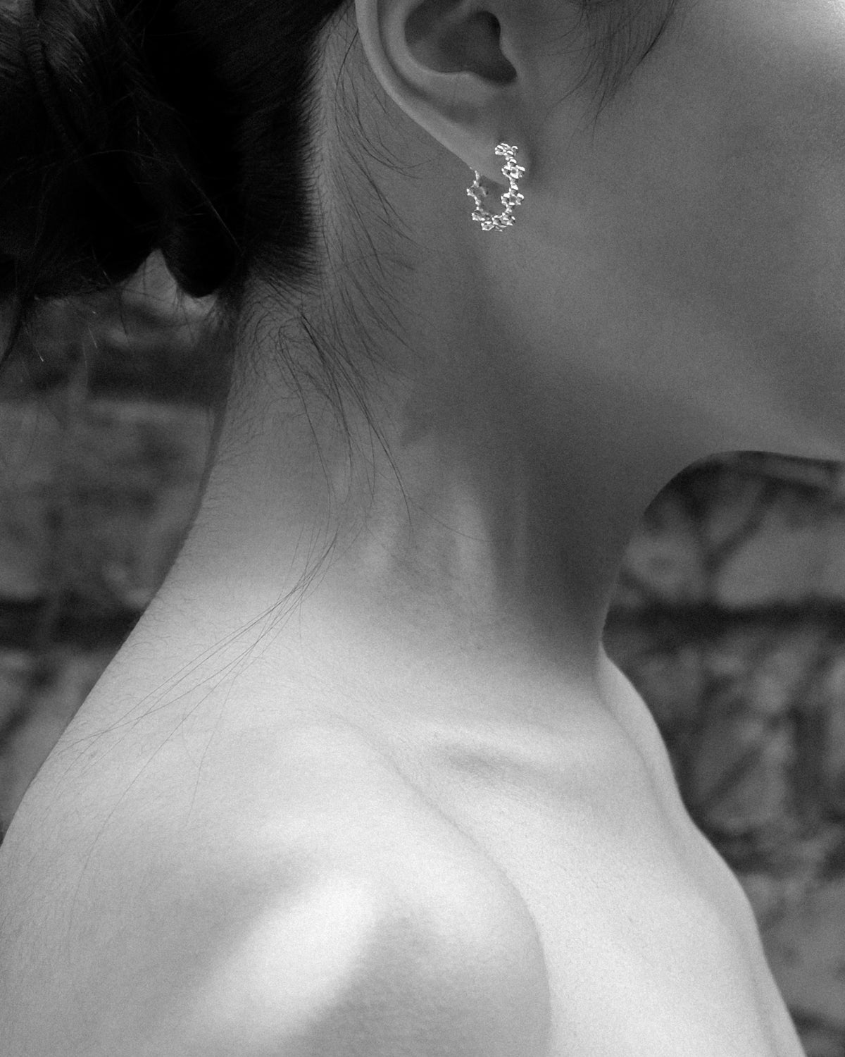 A close-up black and white photograph of a woman’s ear adorned with a delicate silver hoop earring featuring intricate floral details. The sculptural curves of the earring embrace the ear gracefully, highlighting the craftsmanship and organic inspiration behind the design. Soft monochrome lighting captures the subtle texture of the skin, the refined shine of the silver, and the timeless elegance of nature-inspired jewellery.