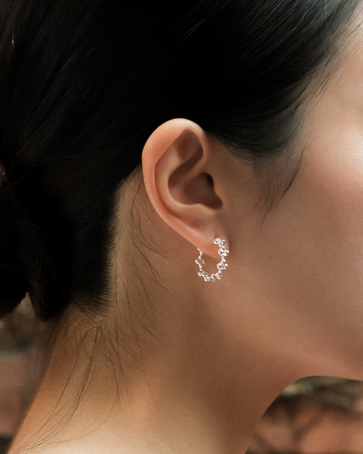 A close-up photograph of a woman’s ear adorned with delicate silver floral details and a silver sphere hoop earring, set against a textured brick wall with soft green vines in the background. The organic floral elements of the jewellery blend with the surrounding nature, creating a refined contrast between handcrafted silver, architectural texture, and quiet natural beauty.