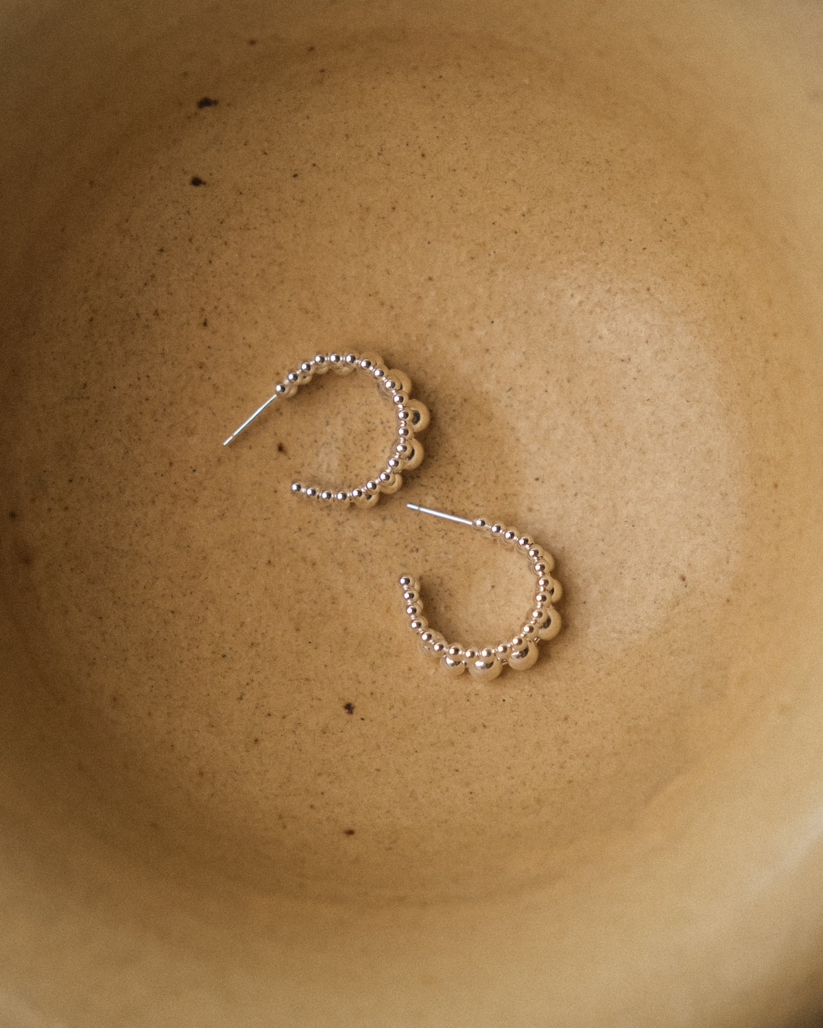 A pair of silver flowing ball huggie hoop earrings with scalloped edges and beaded details, resting inside a textured beige ceramic bowl.