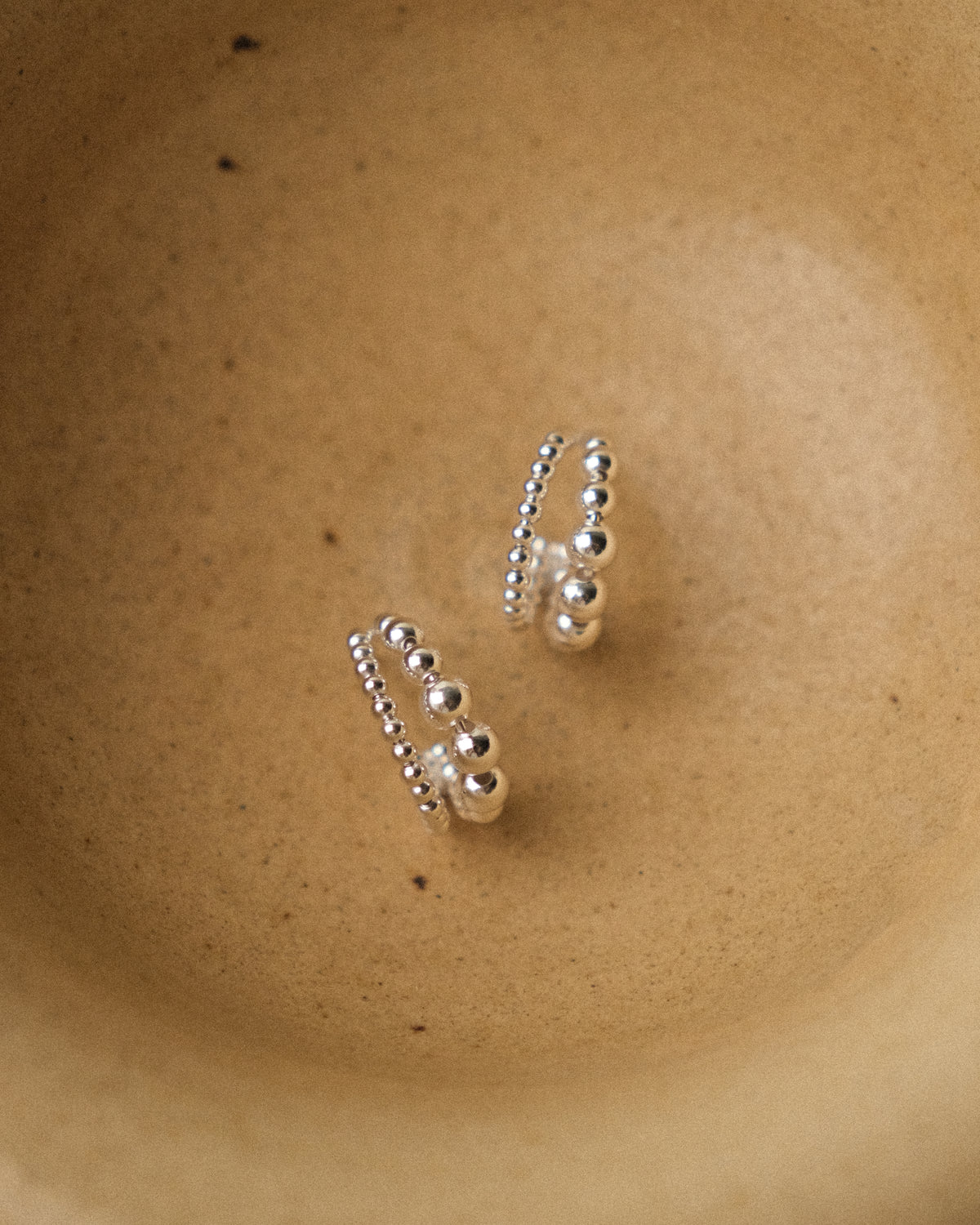 A pair of silver flowing ball hoop earrings with big and small bead details, resting on a textured beige ceramic surface.
