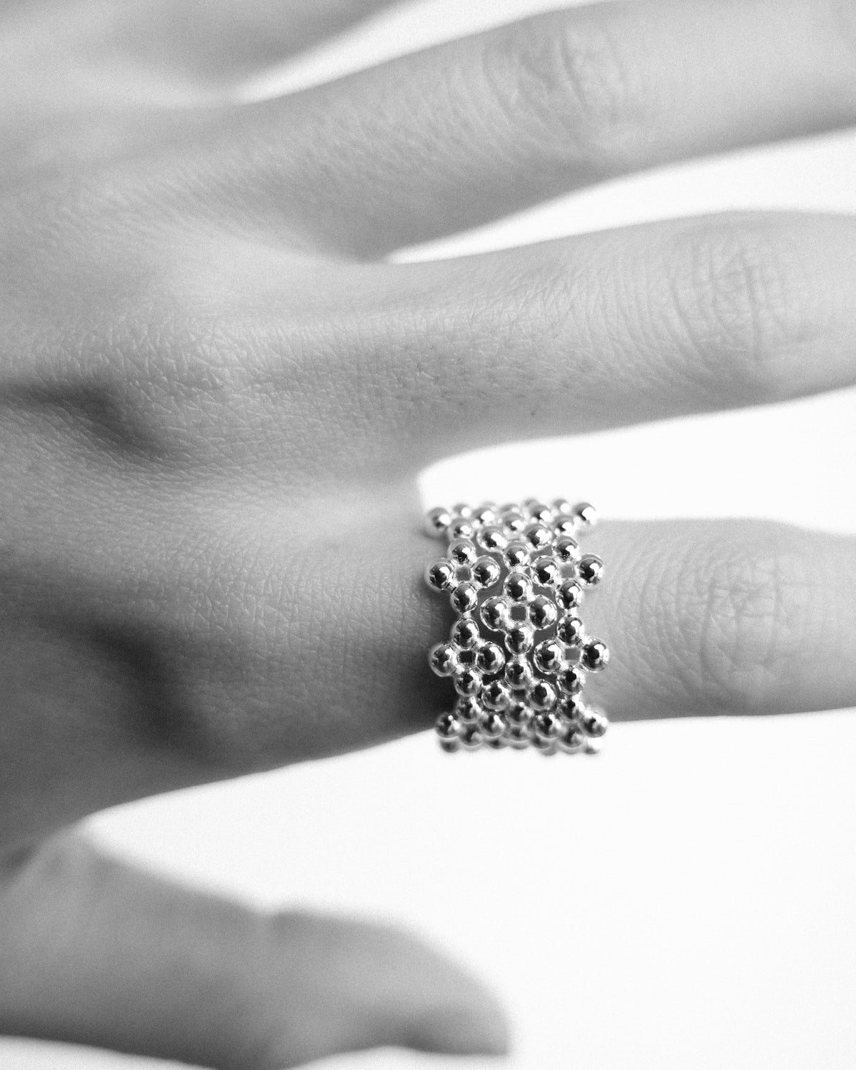 A close-up black and white photograph of a hand adorned with a stack of three delicate silver floral-shaped rings on the index finger. The layered rings create a sculptural composition, highlighting the organic floral forms and handcrafted details of the silver. Soft monochrome tones reveal the texture of the skin, the subtle shine of the metal, and the timeless elegance of minimalist jewellery styling.