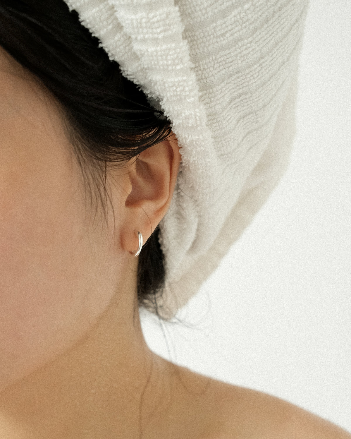 A close-up, an ear adorned with a silver hoop, hair wrapped in a white towel.