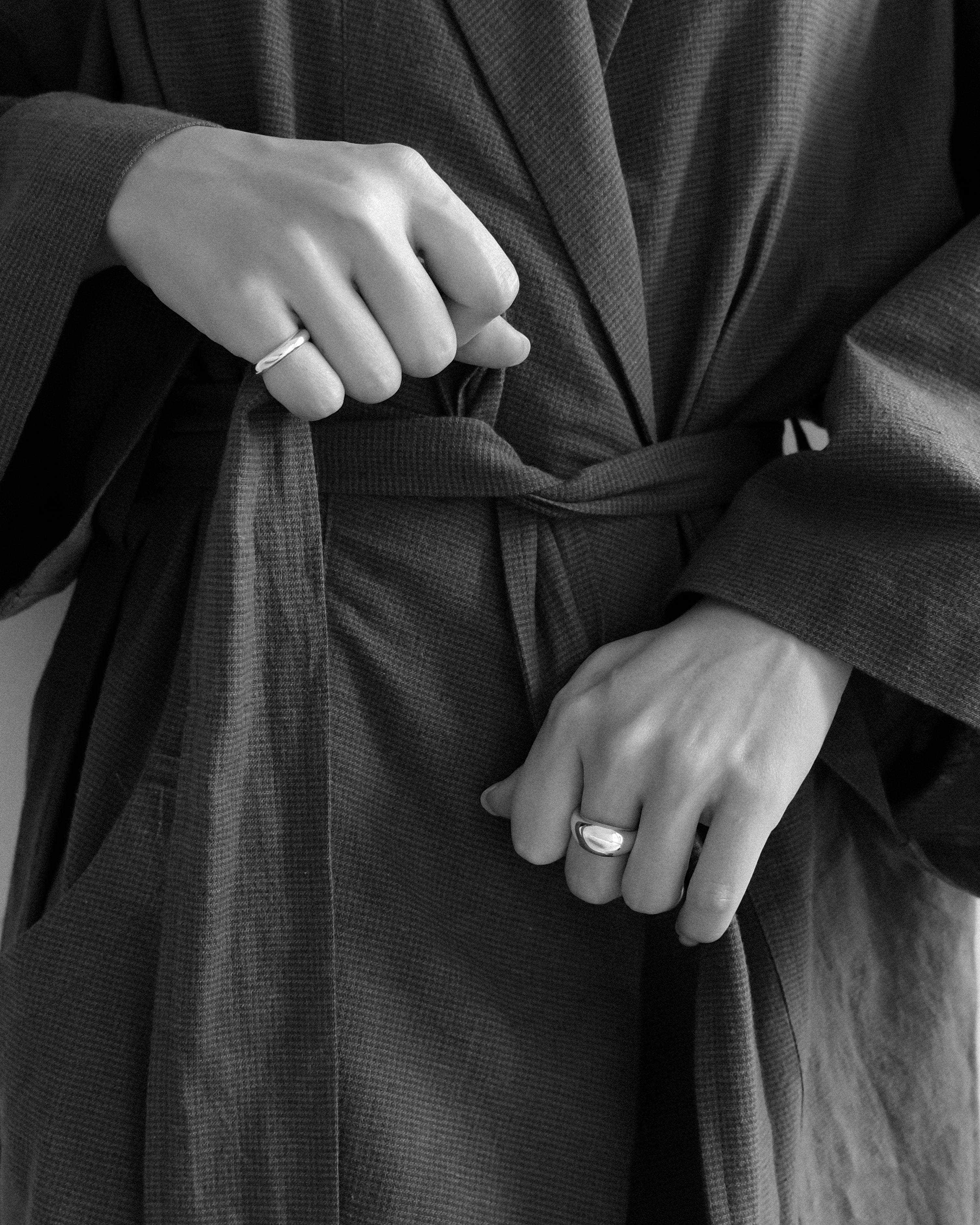 A person in a soft bathrobe, gently adjusting the belt, their hands revealing a bold egg dome ring on the left hand and a small round donut ring resting on the right pinky.