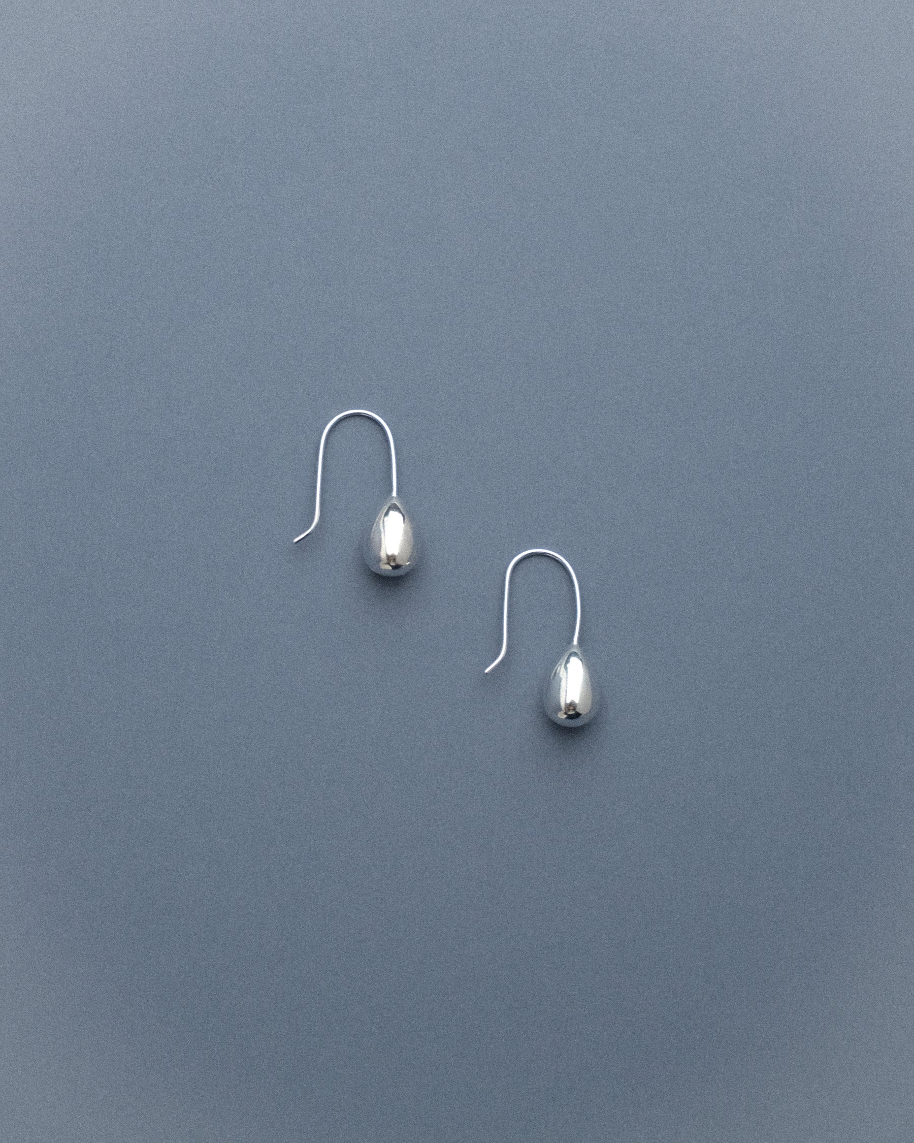 A pair of silver egg drop dangle earrings captured from the side, resting against a soft grey-blue background, their sculptural curves catching the light gently.