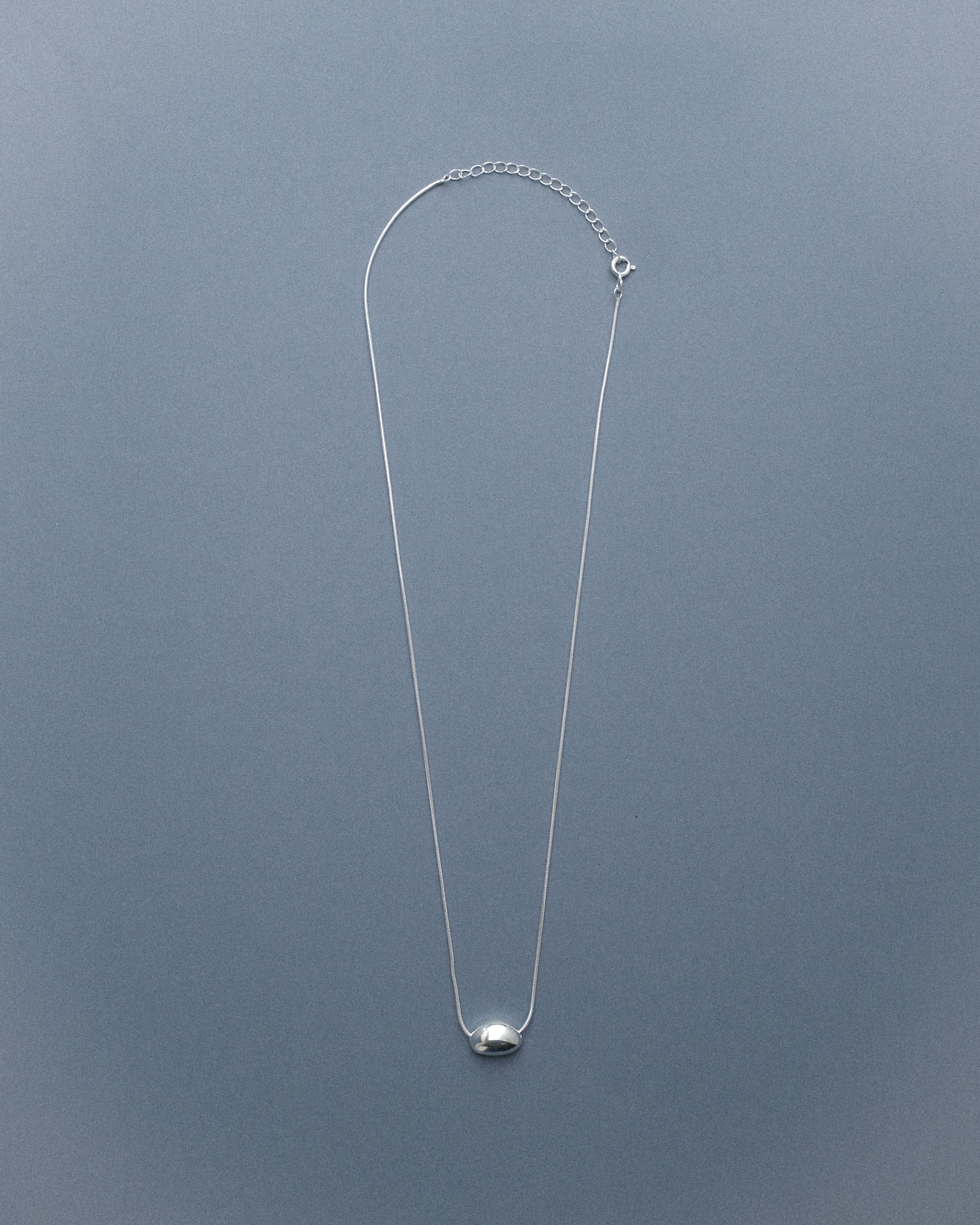 A silver egg-form necklace displayed against a soft geay-blue background, highlighting its smooth, sculptural silhouette.