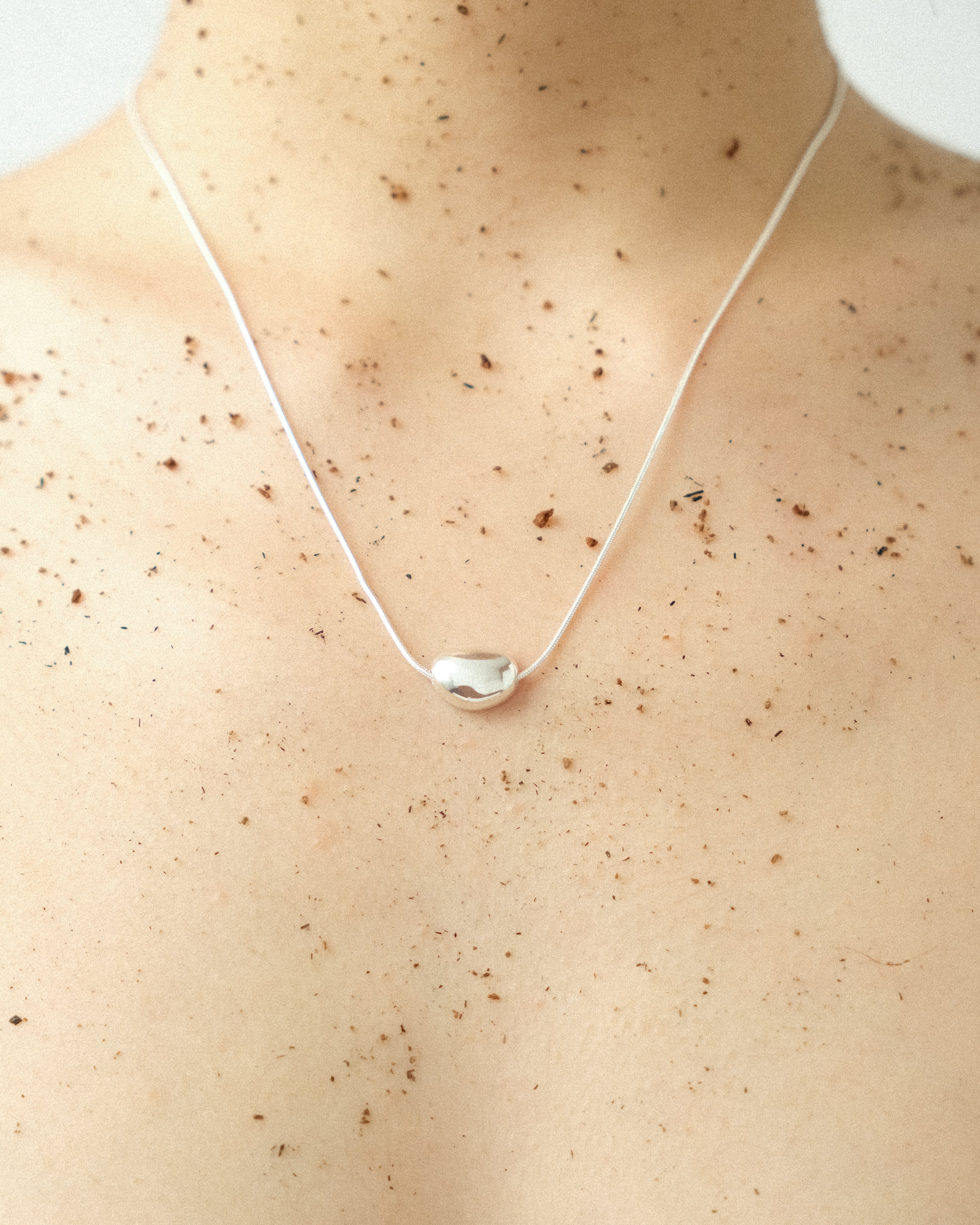 A woman wears a silver egg-form necklace, her skin lightly dusted with soil, set against a crisp white background.