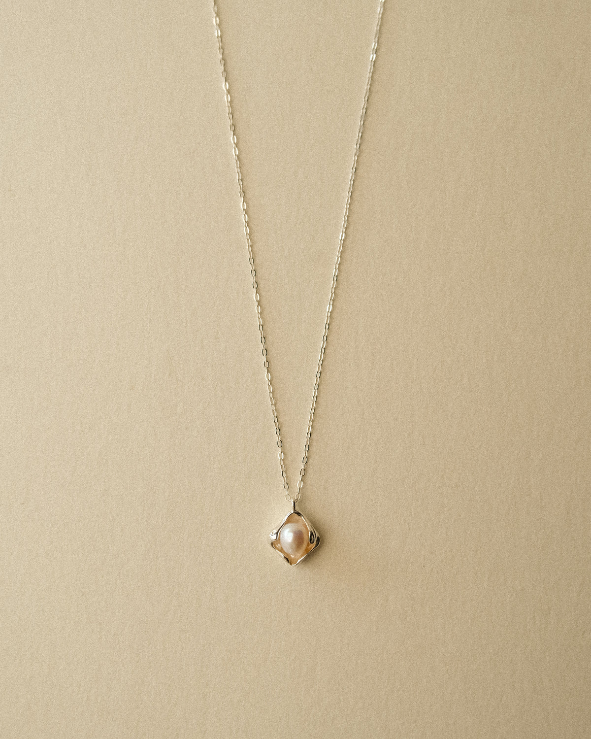 A close-up of a delicate silver chain necklace with a small, petal-shaped pendant featuring a central freshwater pearl, displayed against a warm Wabi-Sabi-inspired background.