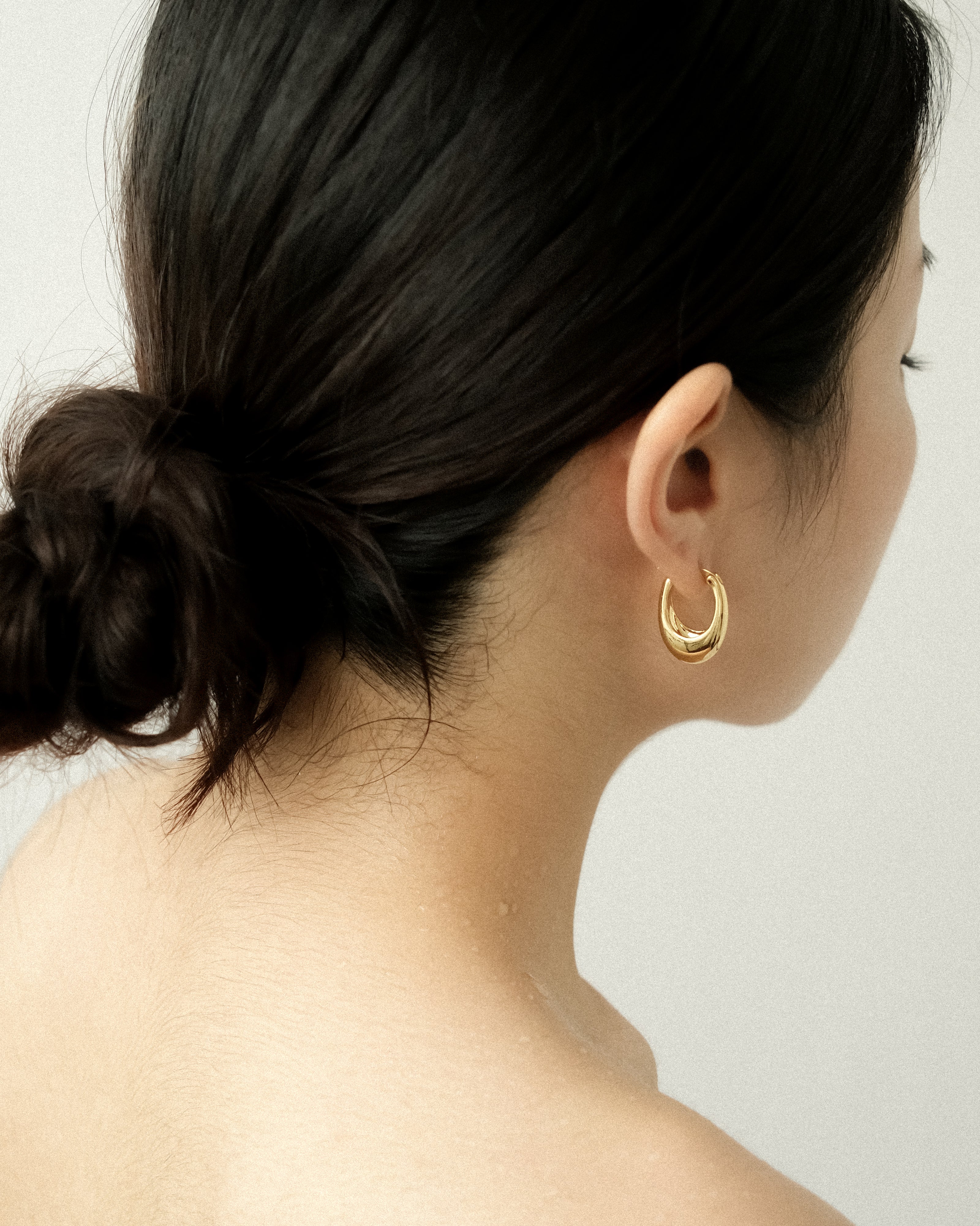 A woman wearing a gold egg drop earring, captured from the side, her hair tied in a neat bun against a clean white background.