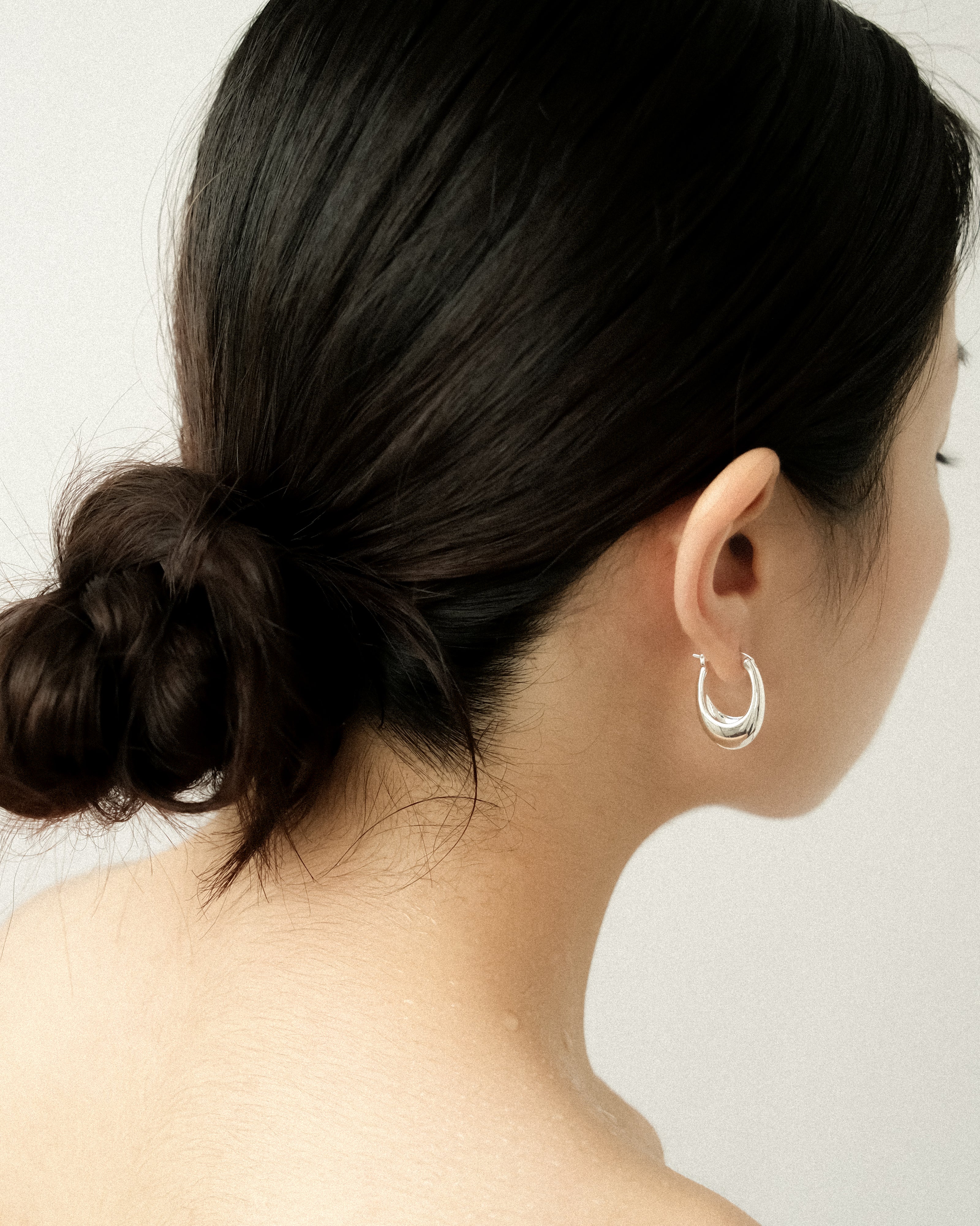 A woman wearing a silver egg drop earring, captured from the side, her hair tied in a neat bun against a clean white background.