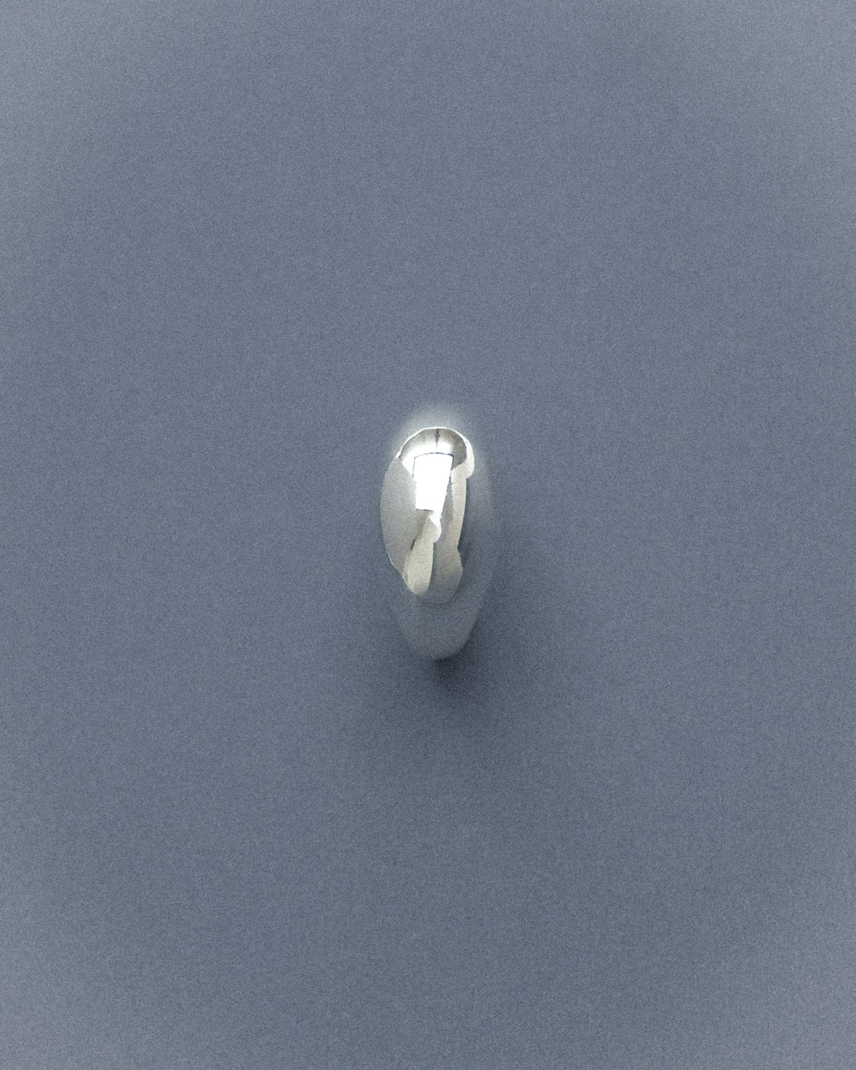 A silver ring captured from the front, resting against a muted grey-blue background, its form quietly defined in soft, understated light.