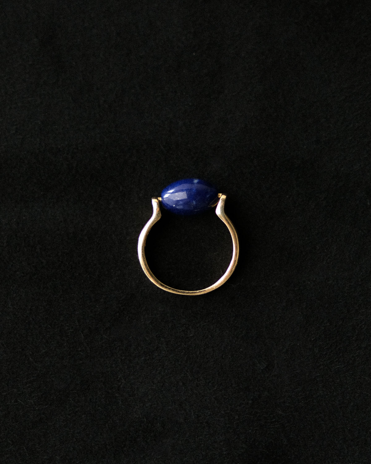 A gold ring featuring a smooth, oval-cut dark blue lapis lazuli stone, presented on a textured black background.