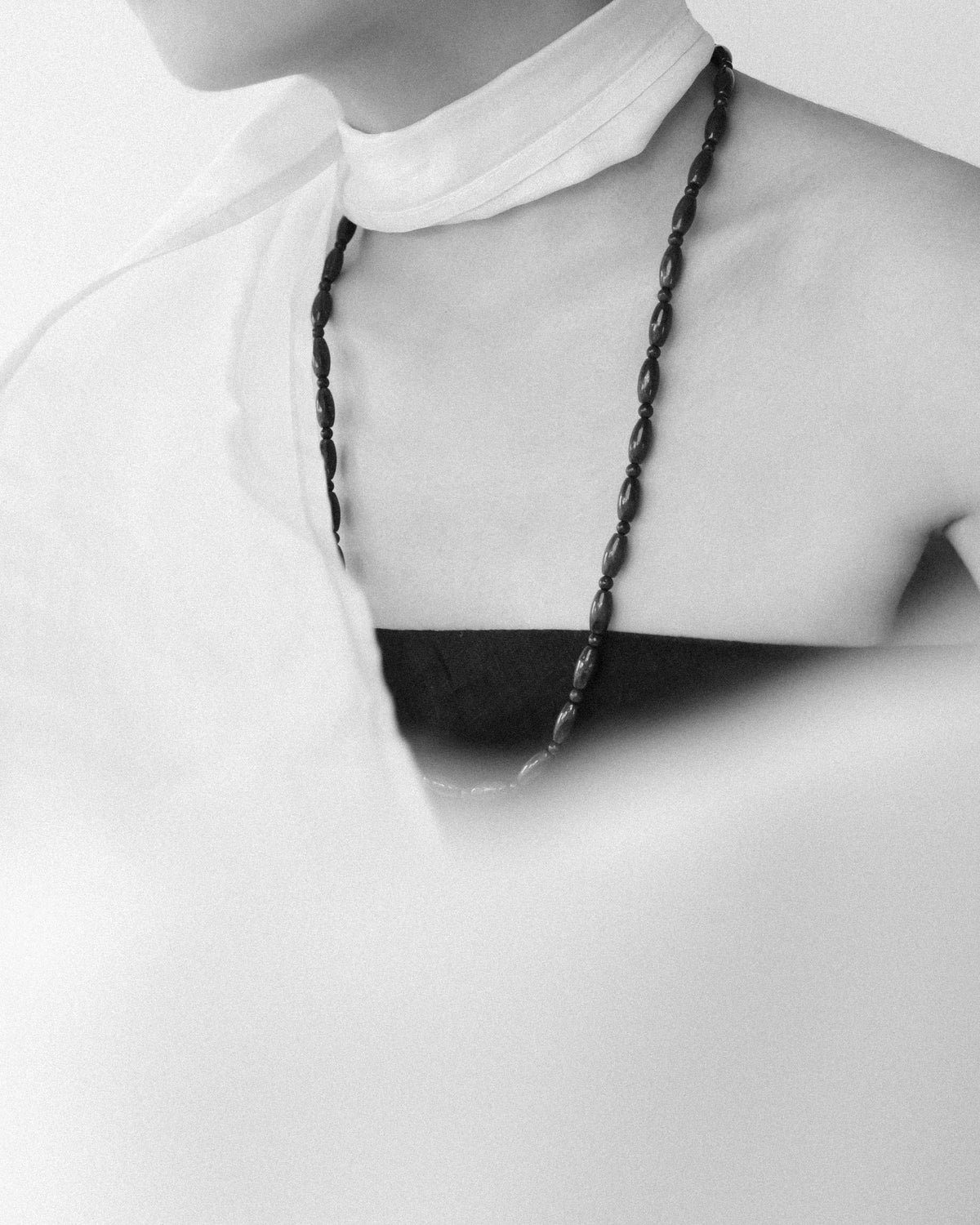 A close-up black and white photograph of a model wearing a long dark blue lapis beaded necklace over a tube dress with a flowing cream white scarf.