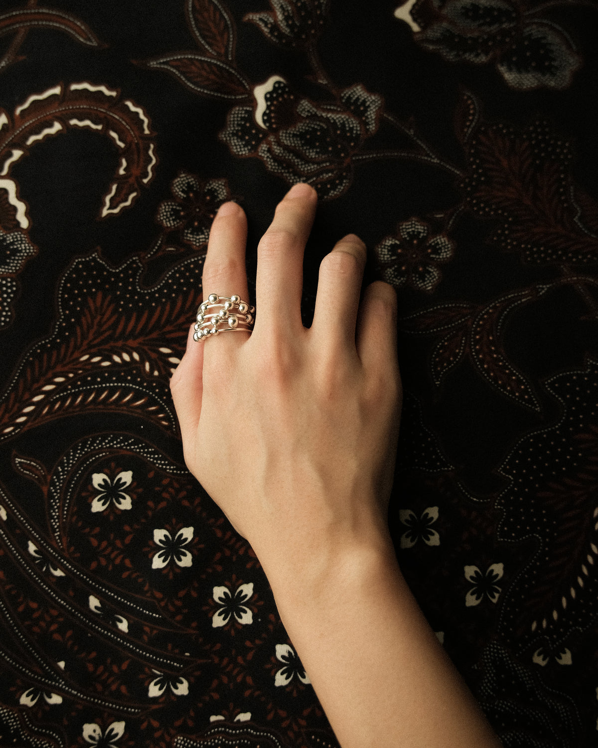 A hand wearing a bold beaded ring on the index finger, set against a deep, dark batik background.