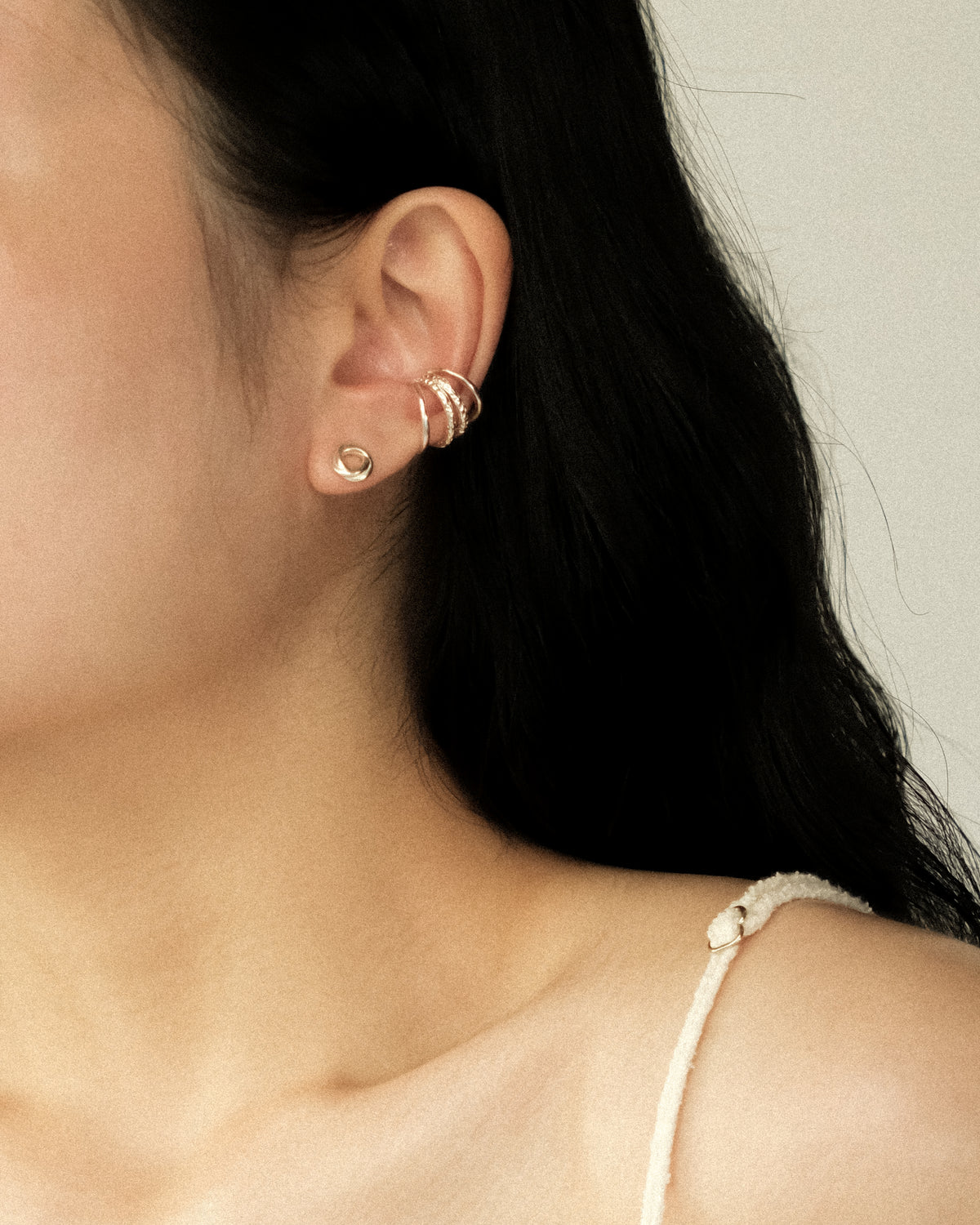 A close-up of a model’s ear wearing a pair of delicate silver-toned ear cuffs, paired with a tiny silver intertwined circle stud, against a soft, warm-toned background.