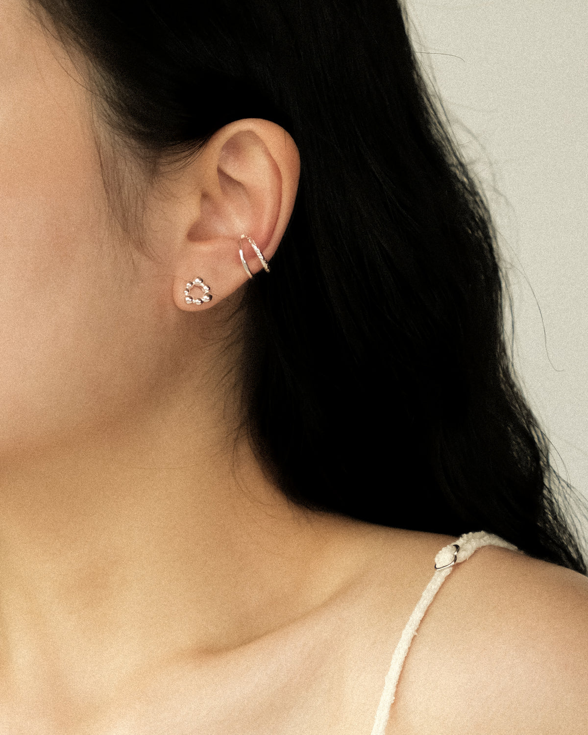 A close-up of a model’s ear wearing a delicate silver-toned ear cuff, paired with a tiny silver irregular sphere stud, against a soft, warm-toned background.