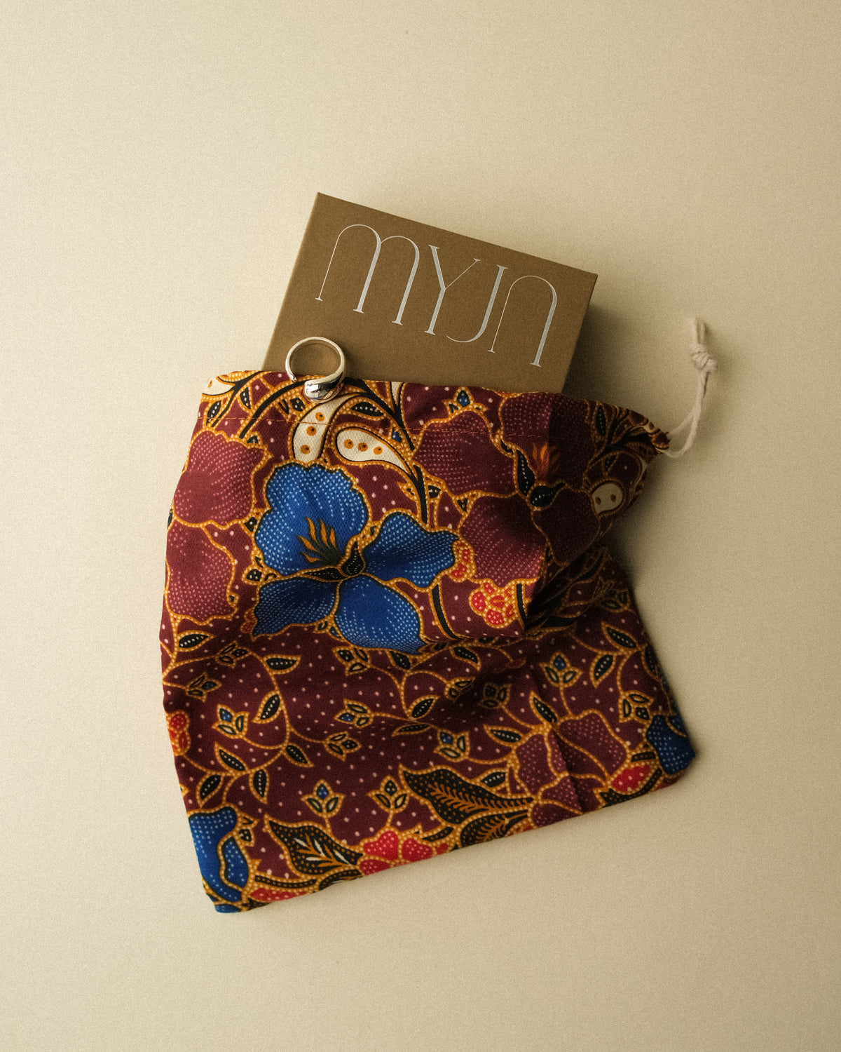 A khaki box with the “MYJN” logo, featuring a colourful batik-inspired floral and paisley pattern in red, blue, yellow, and white, set against a beige background and styled with a silver ring placed on top of the box.