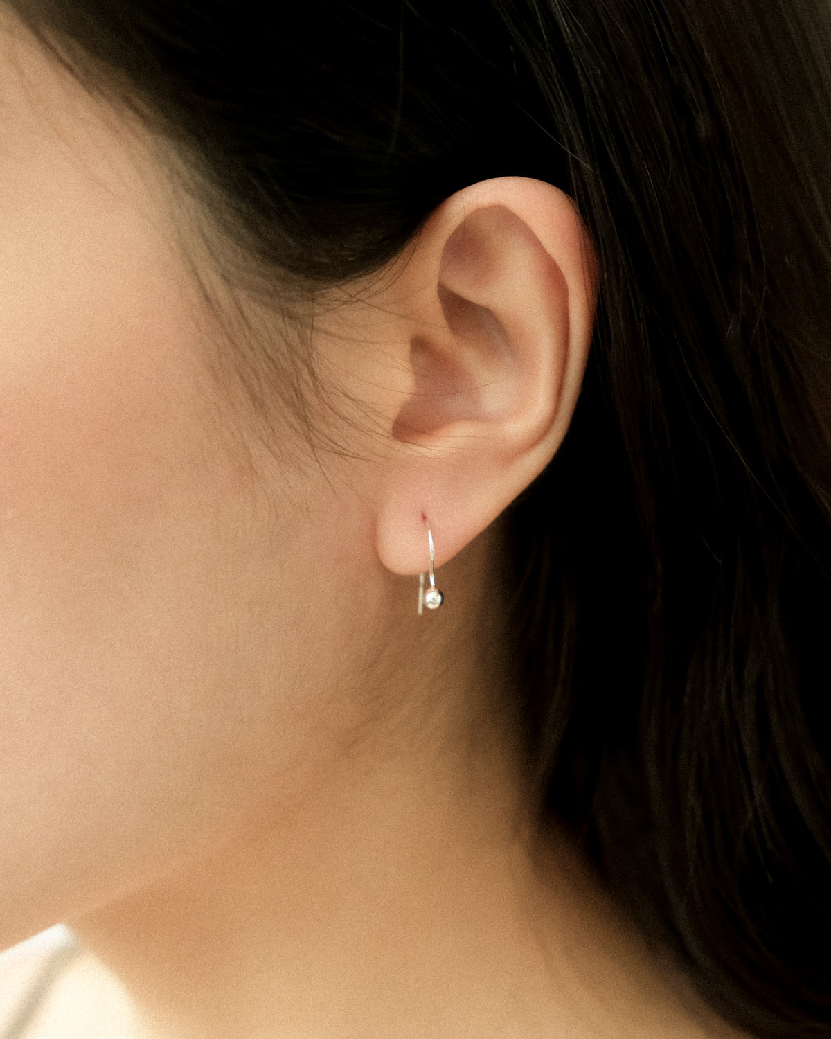 Close-up of an ear wearing a tiny silver dangle ball earring, captured from side.