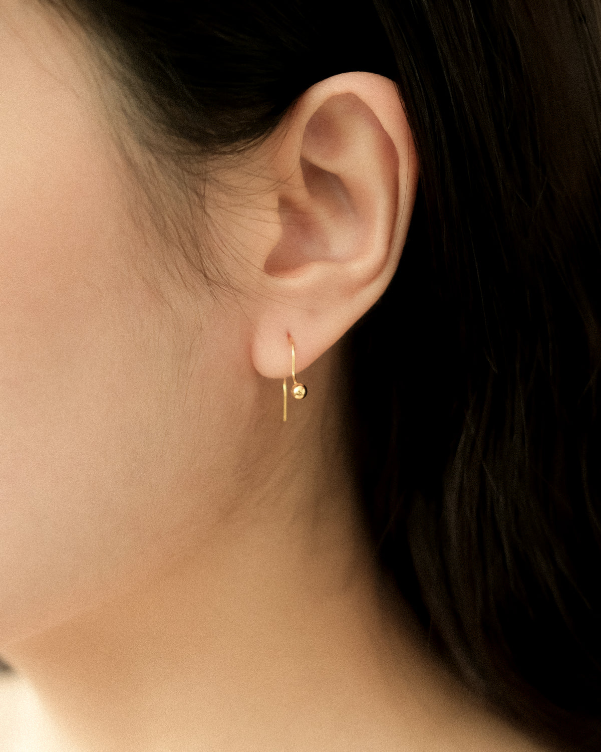 Close-up of an ear wearing a tiny gold dangle ball earring, captured from side.