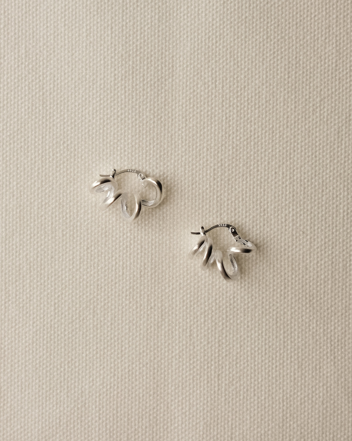 A pair of sculptural curvy sterling silver hoop earrings with a brushed finish, arranged on a textured cream tablecloth.