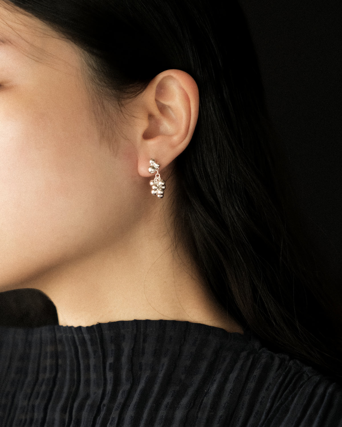 Close up of a model wearing a silver toned grape cluster dangle earring, with polished spheres hanging just below the earlobe against dark hair.