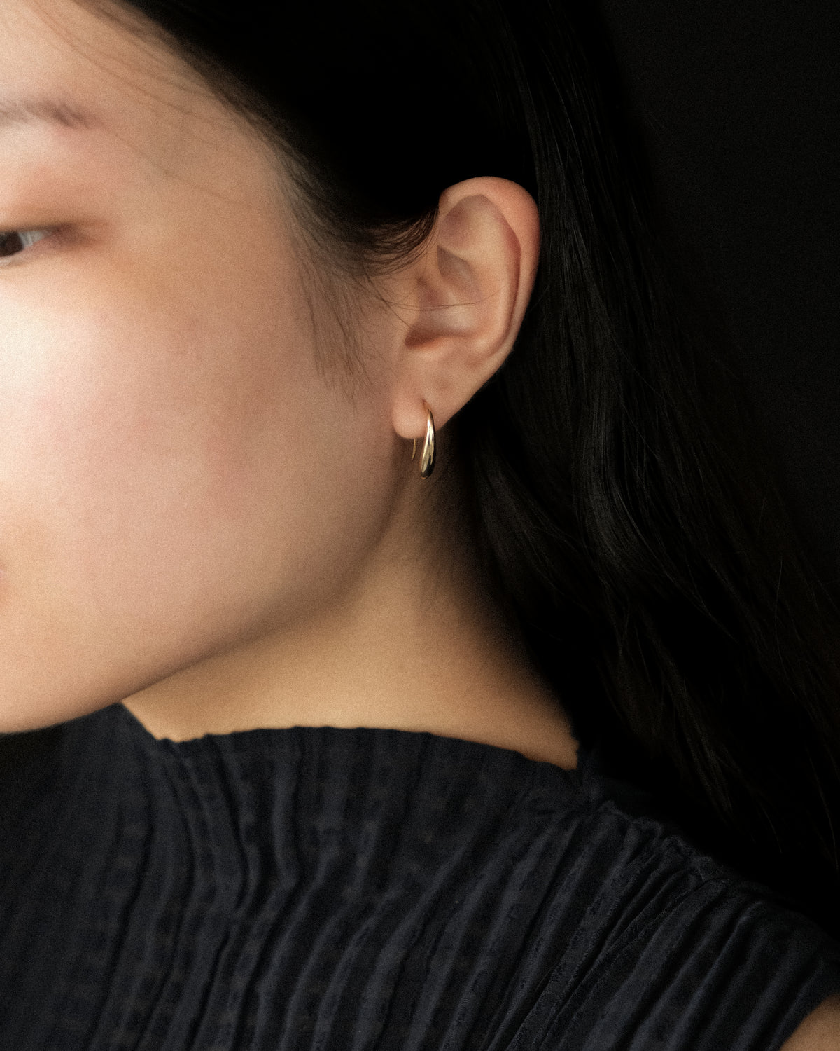 A close-up of a model's ear wearing a small, polished gold hoop earring against a dark background.