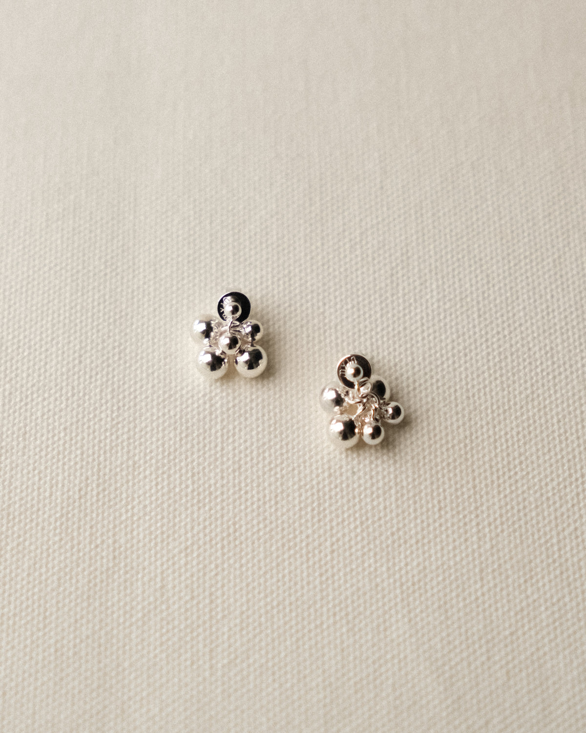 A pair of small silver stud earrings with clustered bead-like grape or geometric designs, displayed on a textured cream fabric background.
