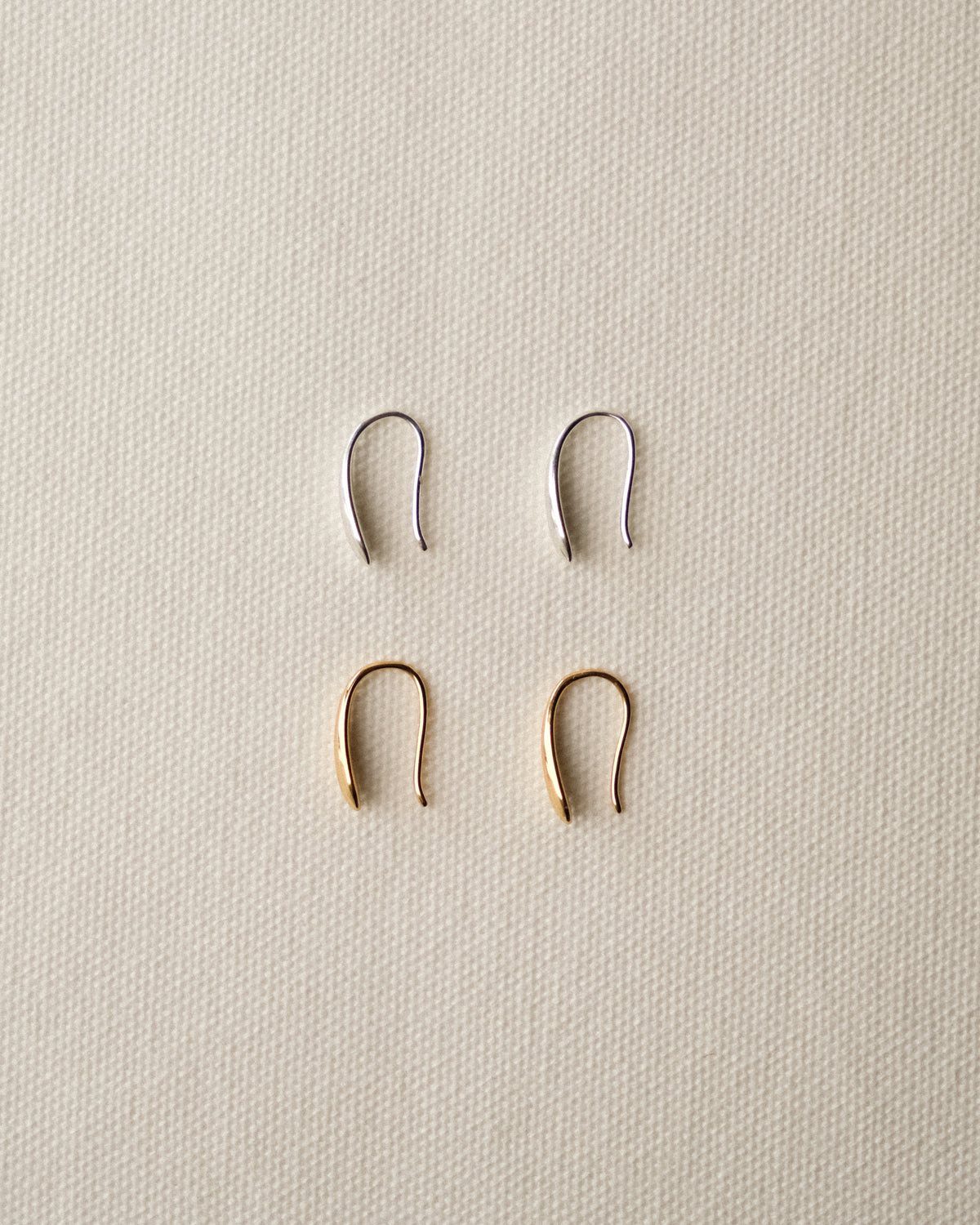 Four small U-shaped metal ear hoops arranged in a 2x2 grid on a textured off-white fabric background. The top row features two silver-toned hoops, and the bottom row features two gold-toned hoops.