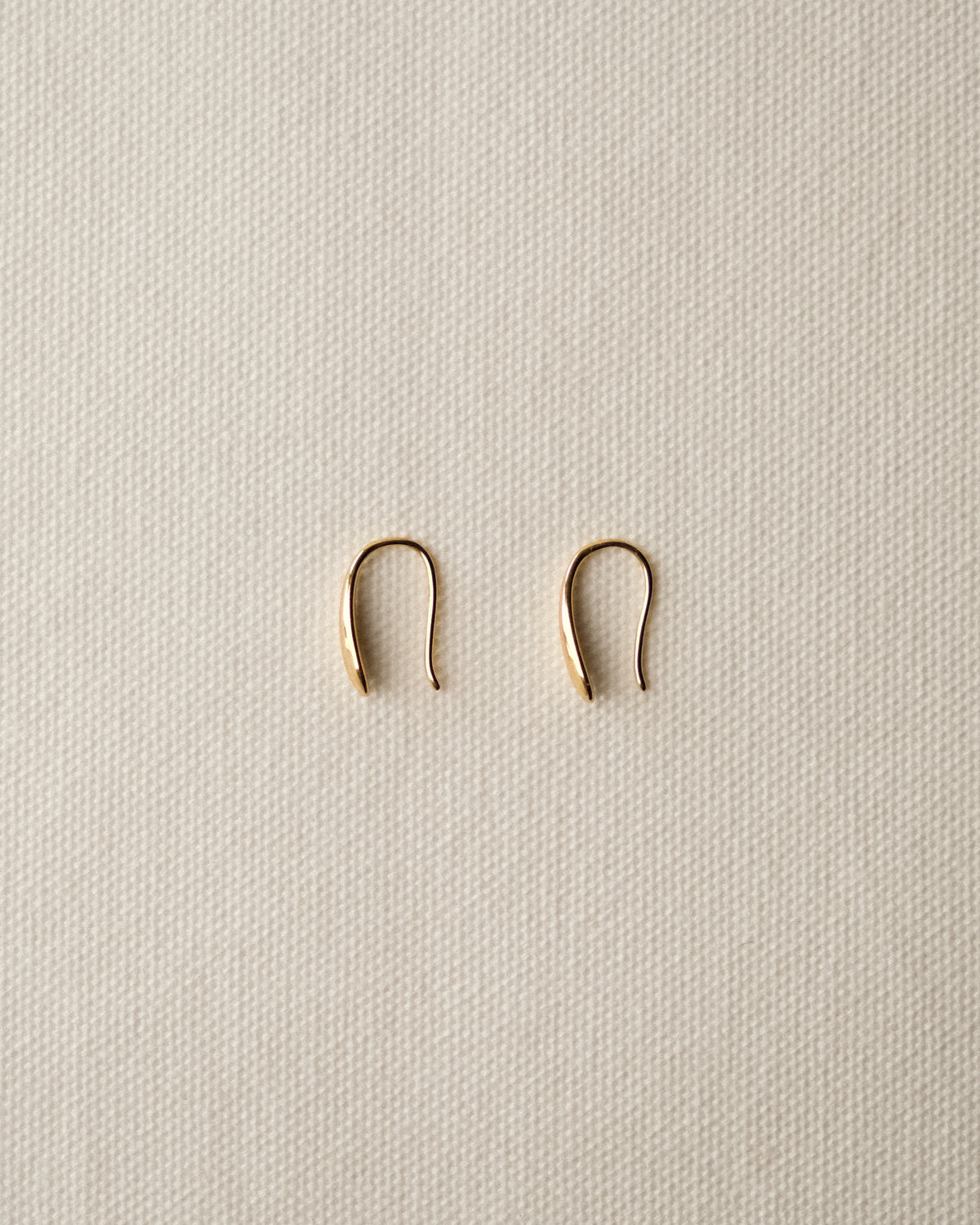 A pair of small gold-toned U-shaped drop earrings displayed on a textured cream fabric background.