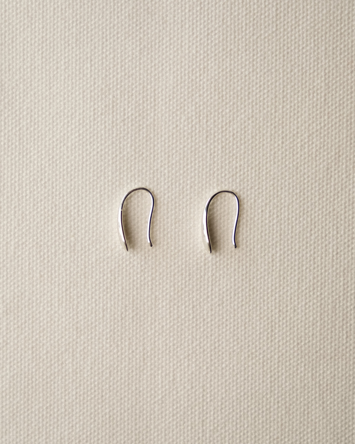 A pair of small silver metal ear hoops, displayed on a textured beige fabric background.