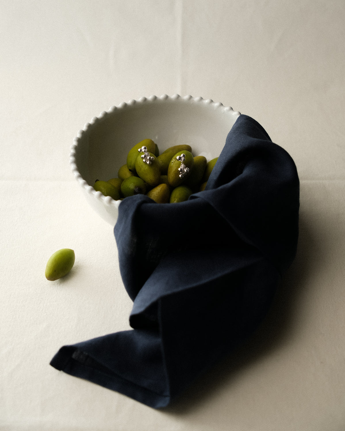 Silver grape cluster dangle earrings resting on green olives in a white scalloped bowl, with navy fabric draped over the rim on a cream tablecloth.