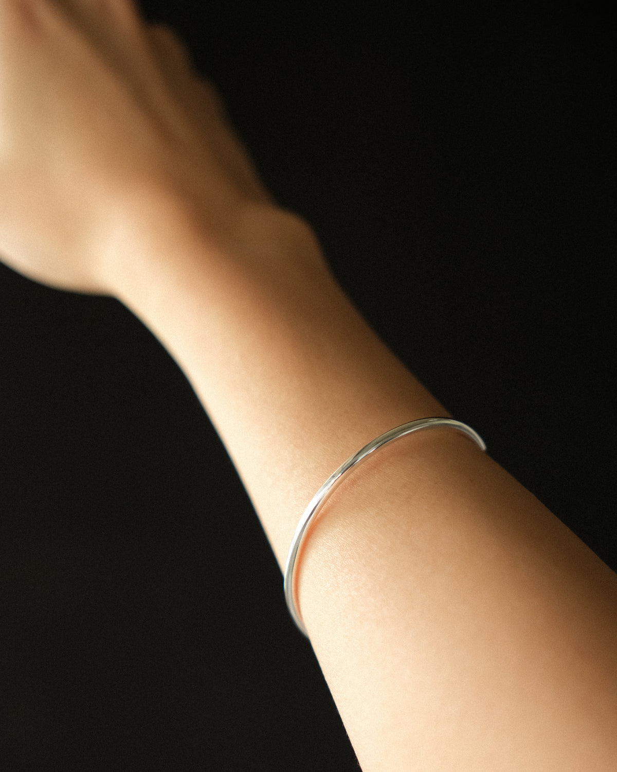 A thin silver polished bangle bracelet worn on a person's wrist against a solid black background.