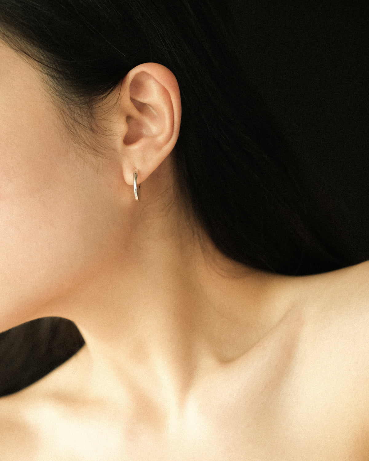 A close-up of a model's ear wearing a delicate, thin silver huggie hoop earring against a dark background.