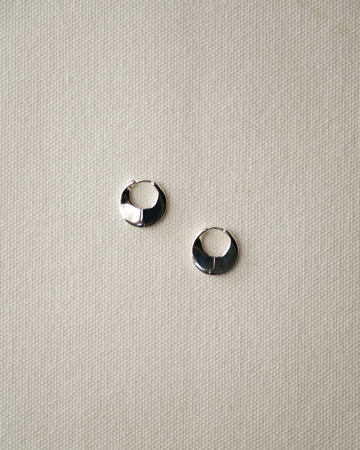 A pair of silver sculptural hoop earrings on a textured beige fabric background.