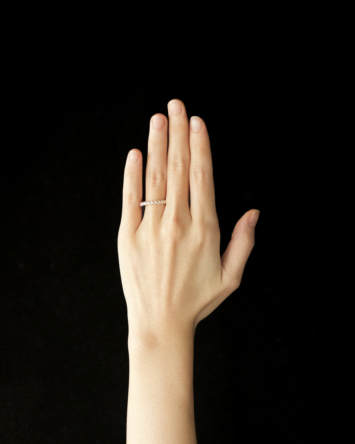 A thin silver twisted band ring worn on the ring finger of a hand, shown against a solid black background.