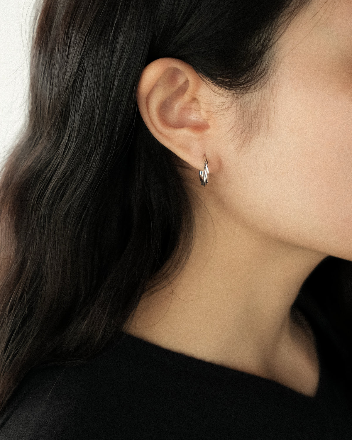A close-up of a woman's ear wearing a small silver twsited hoop earring.