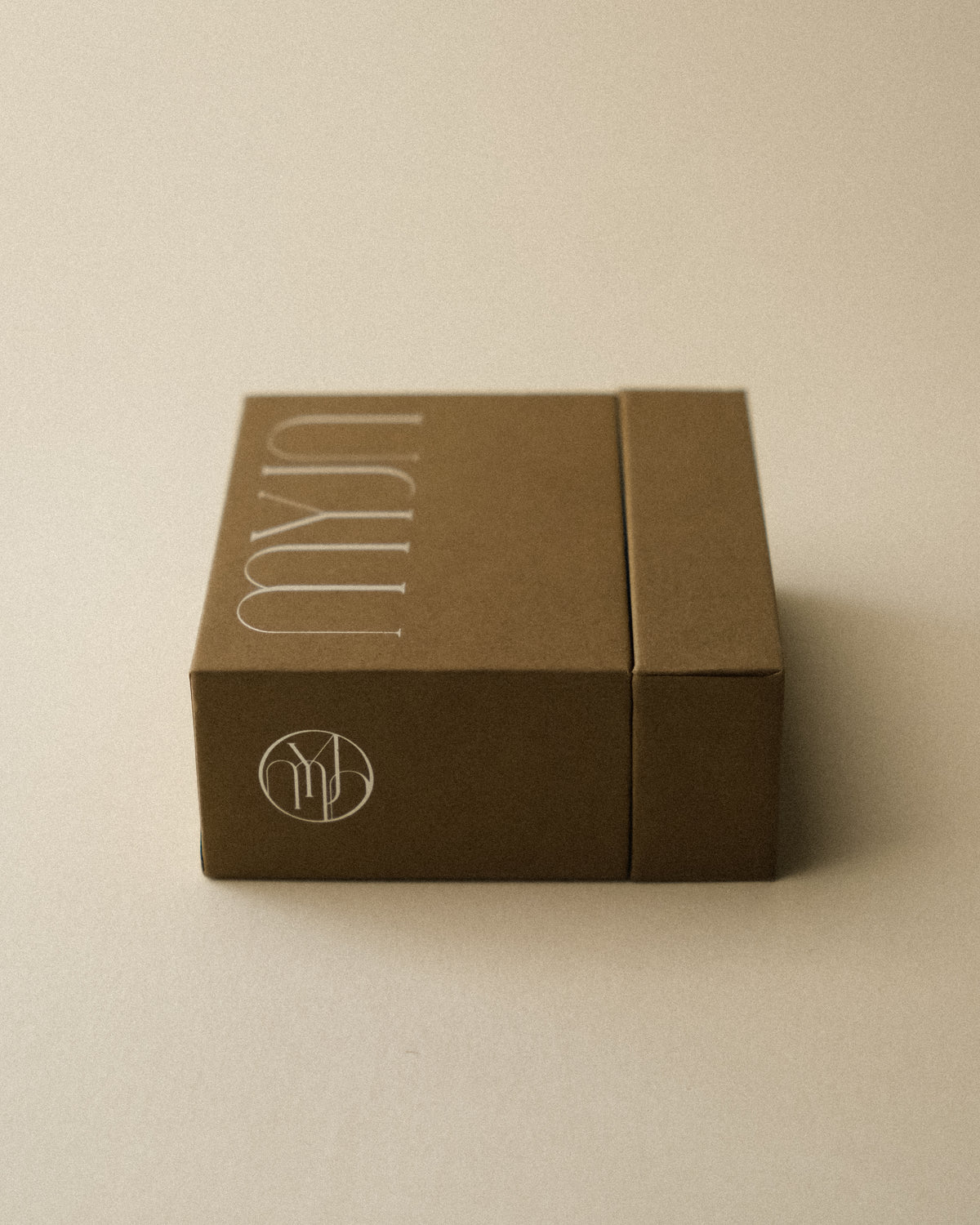 A light khaki brown jewellery box photographed from the side, featuring the MYJN round logo, against a warm toned wabi sabi backdrop.