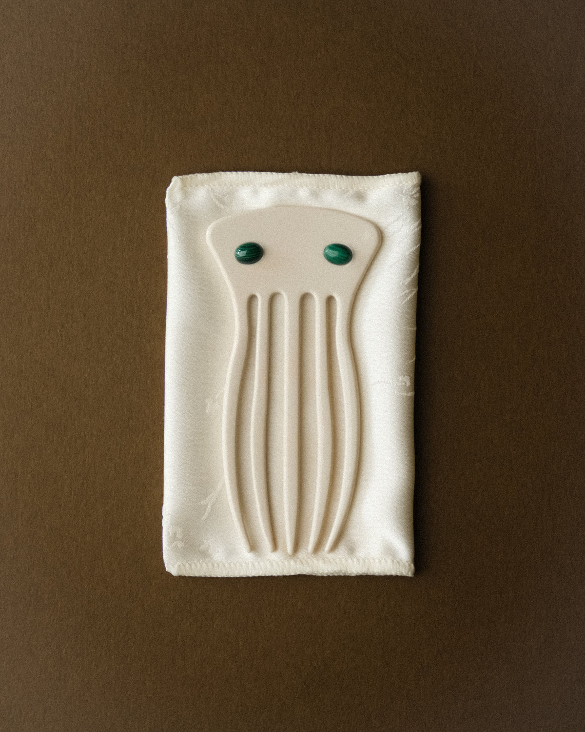 A cream white satin flower print folded pouch and an ivory decorative hair comb adorned with two malachite stones, stacked together against a warm brown wabi sabi backdrop. Soft diffused lighting accentuates the satin's delicate texture, the comb's sculptural silhouette, and the rich green malachite accents, creating a minimal, refined composition with a quiet luxury feel.